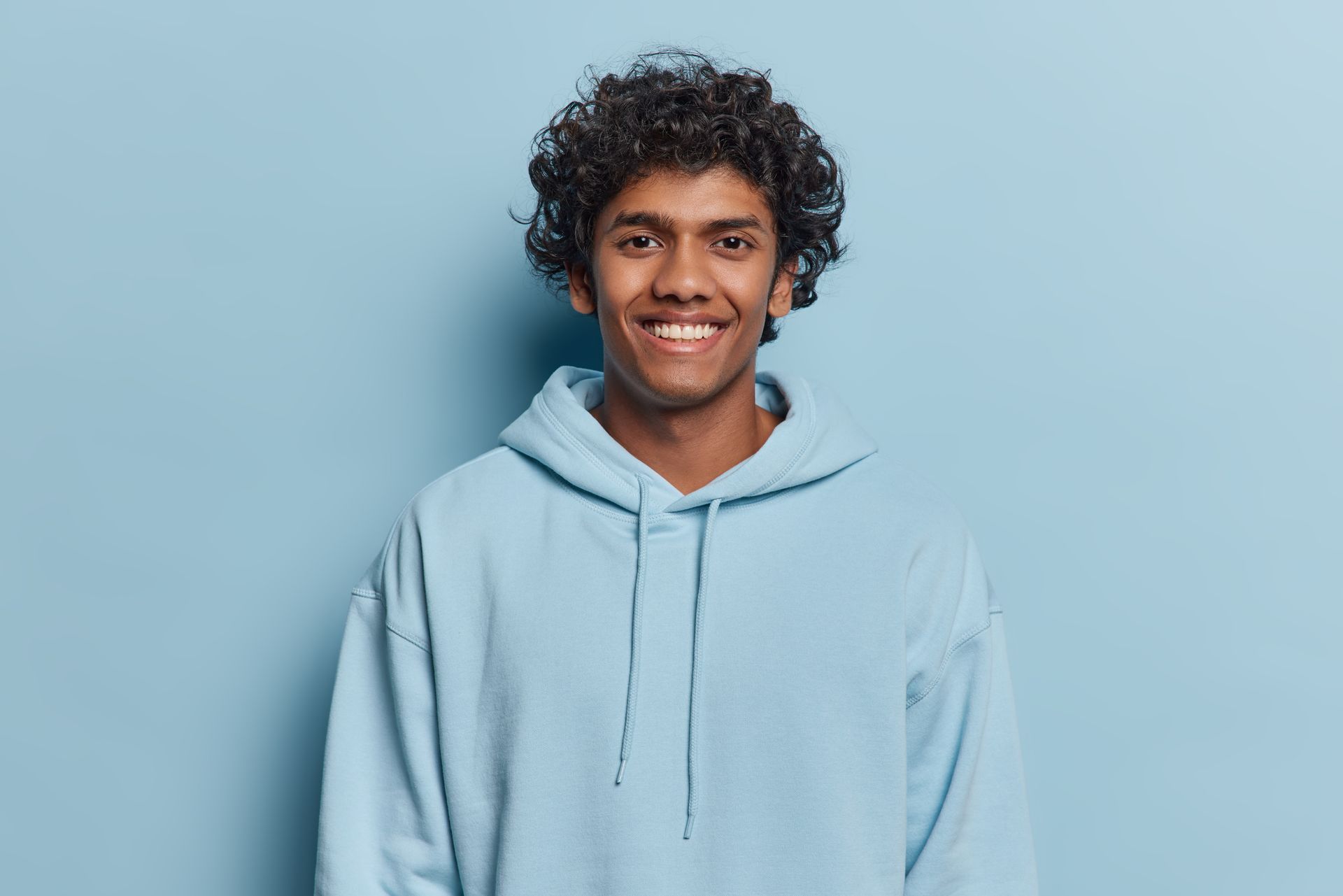 A young man with curly hair is wearing a blue hoodie and smiling.