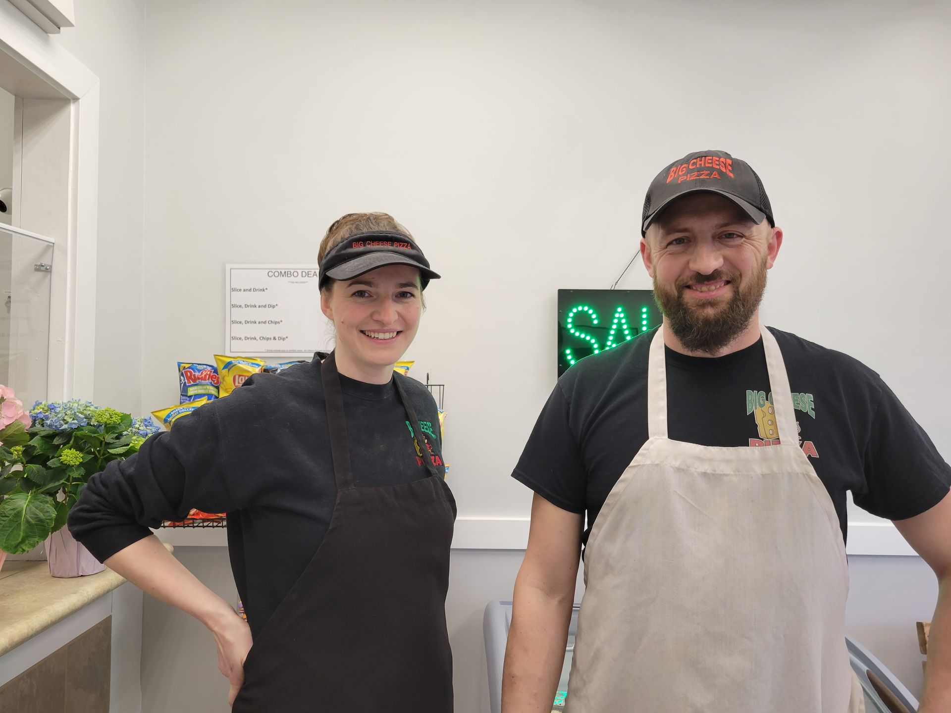 Two people, one in an apron and the other in a visor, stand behind a counter in a shop with a "SALE" sign.