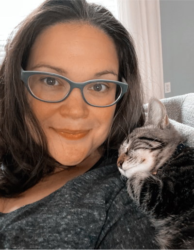 Woman with glasses smiles, cat asleep on her shoulder.