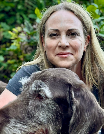 Woman with long blonde hair, smiling, and hugging a brown dog outdoors.