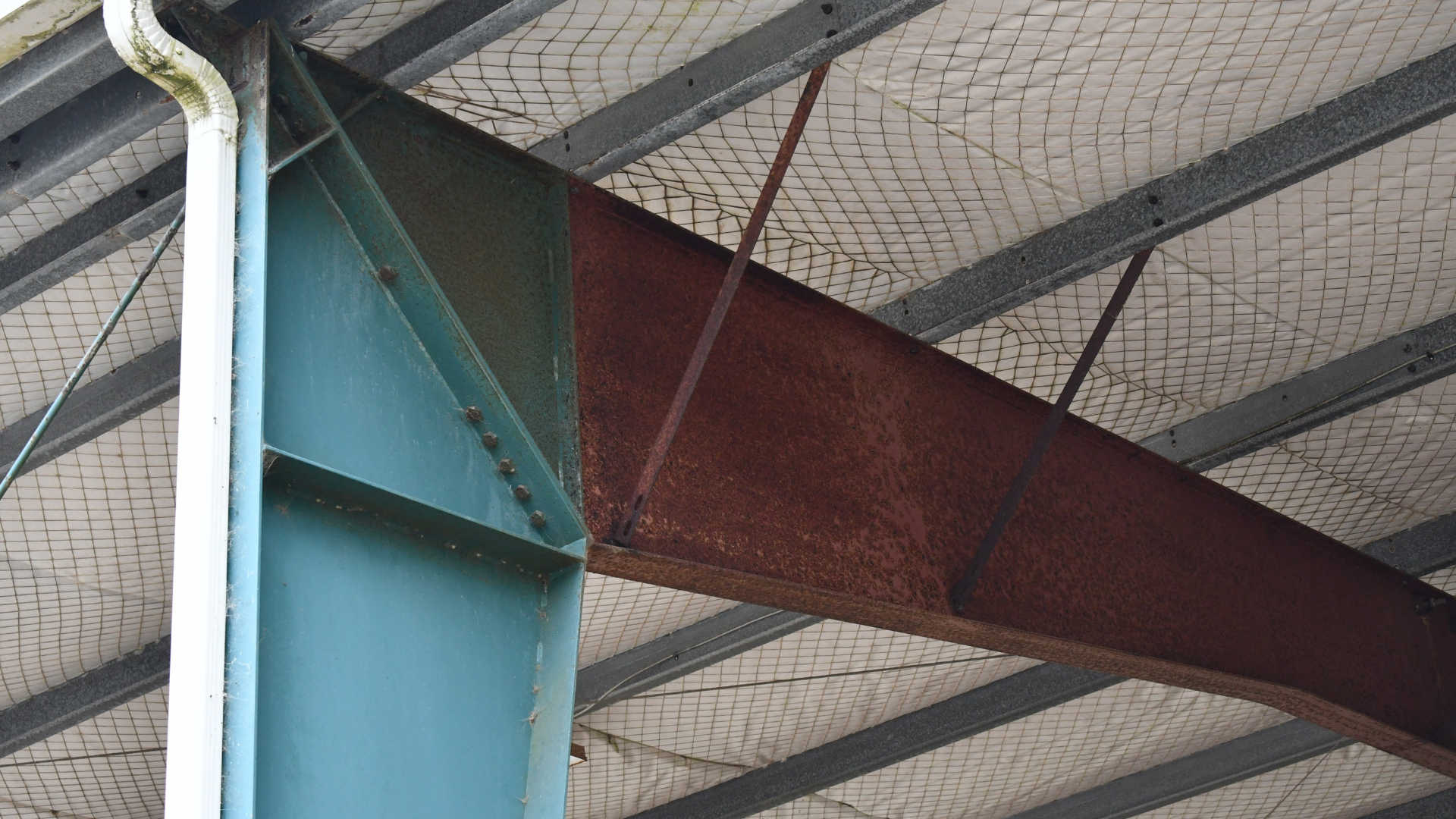 A steel structure's interior view showing a green beam, a rusty brown beam, and a white, textured ceiling.