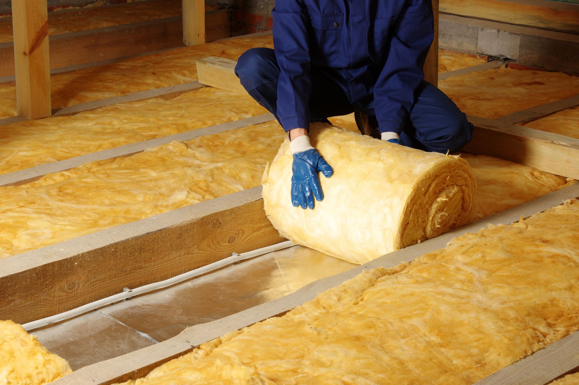 Person installing insulation in an attic, using a roll of yellow insulation between wooden beams.