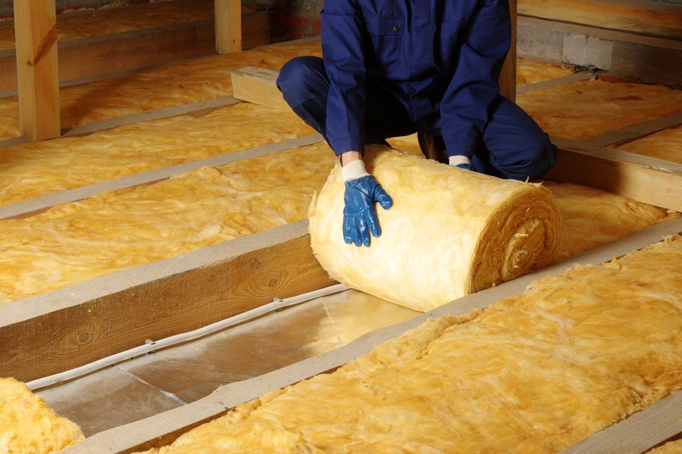Person installing insulation in an attic, using a roll of yellow insulation between wooden beams.