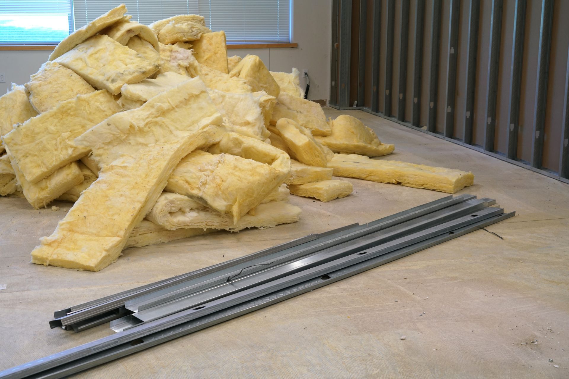 Pile of yellow insulation and metal tracks on a room's concrete floor; next to a wall with vertical grooves.