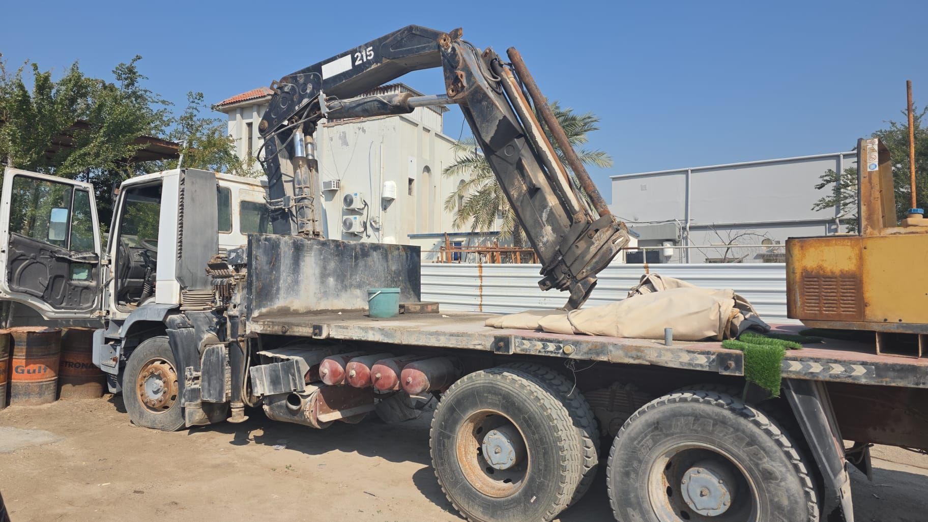 A crane truck with its arm raised. The truck is dirty with a flatbed in a construction area on a sunny day.