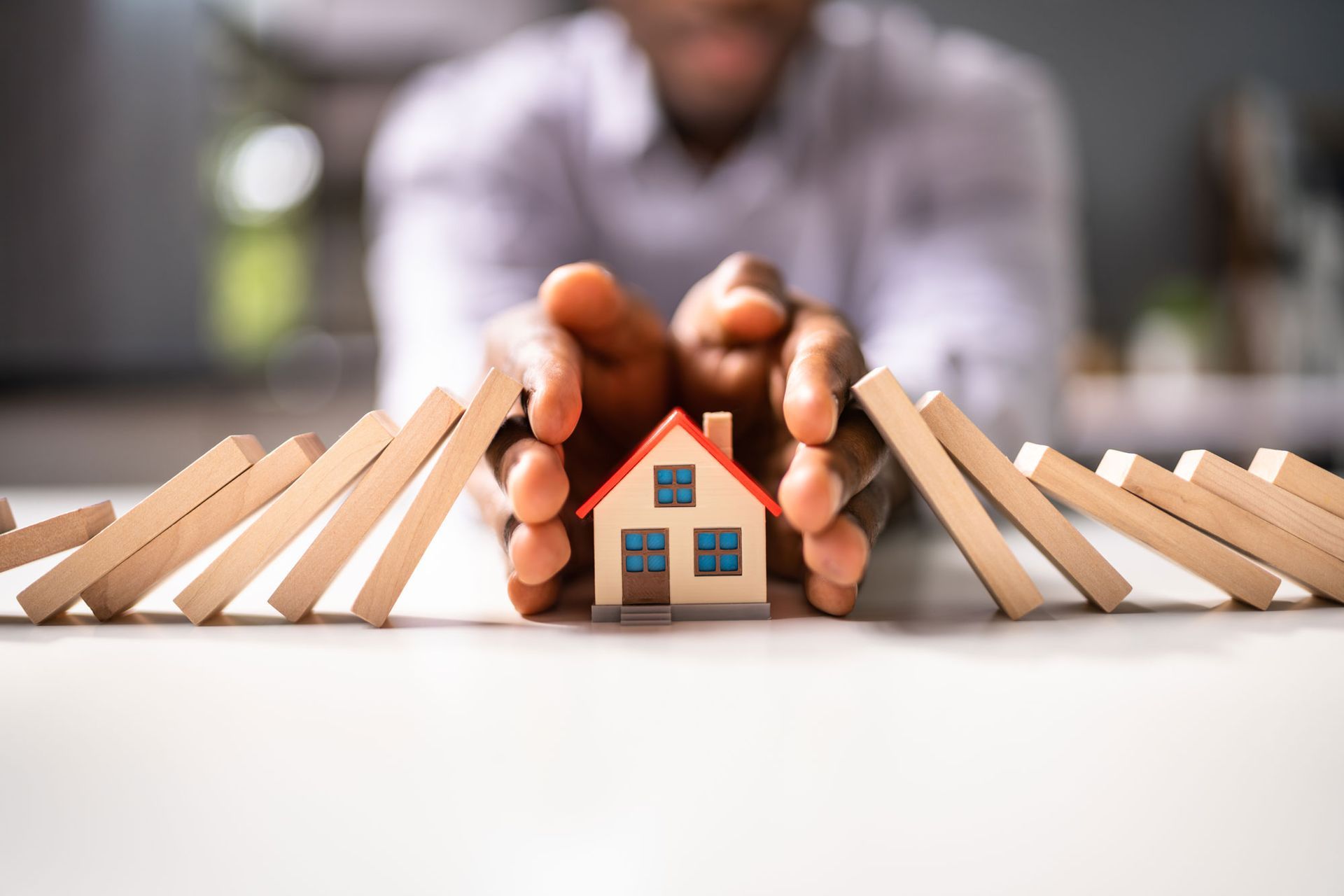 A man is holding a small house in his hands to stop dominoes from falling on it.