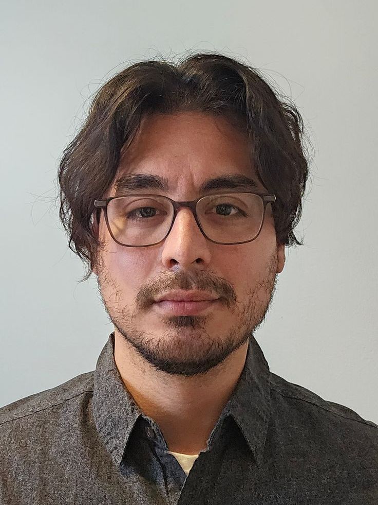 A man with glasses and a beard is standing in front of a white wall.