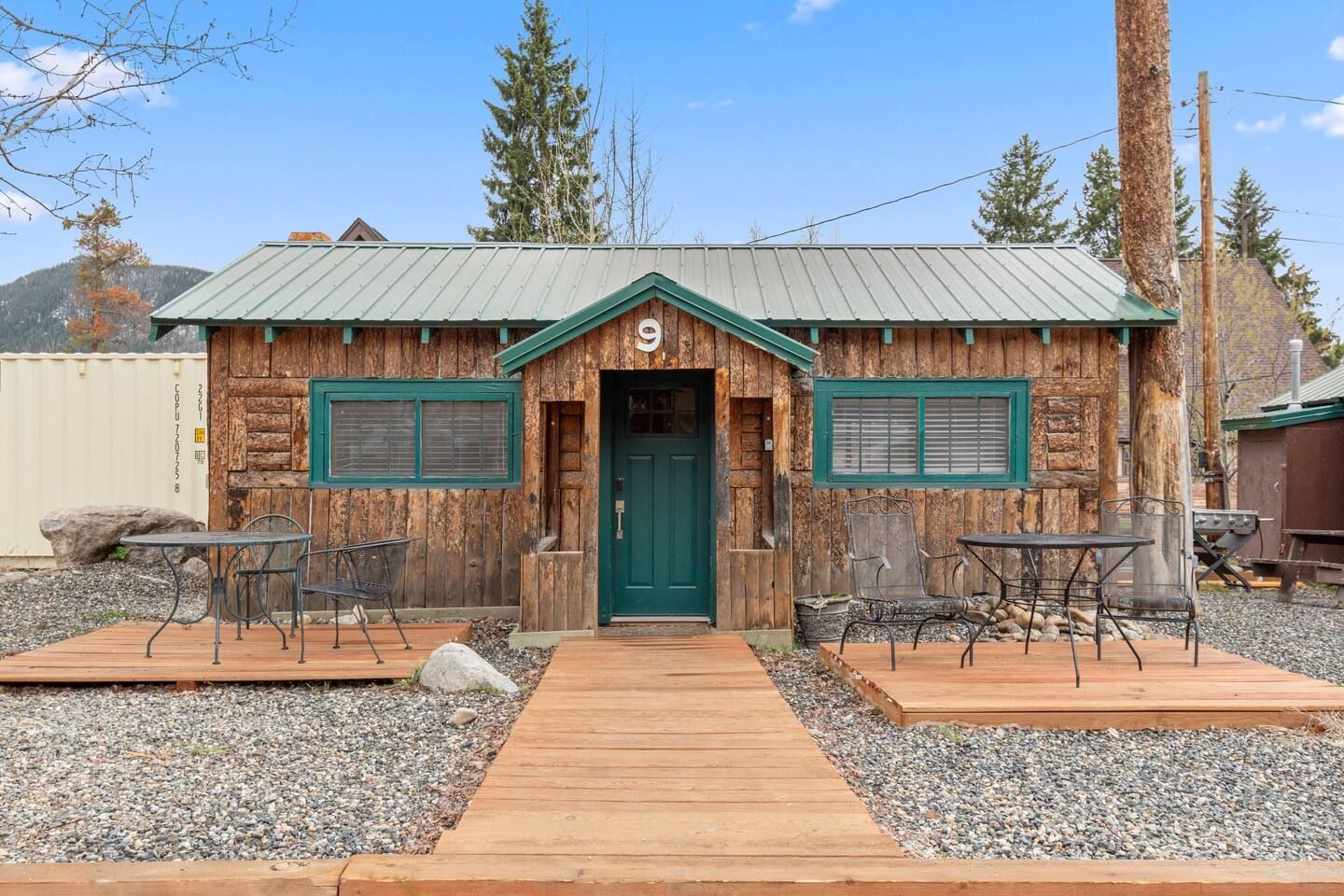 Cabin with green door, windows, and trim. Two small tables and wooden walkway. Gravel yard.