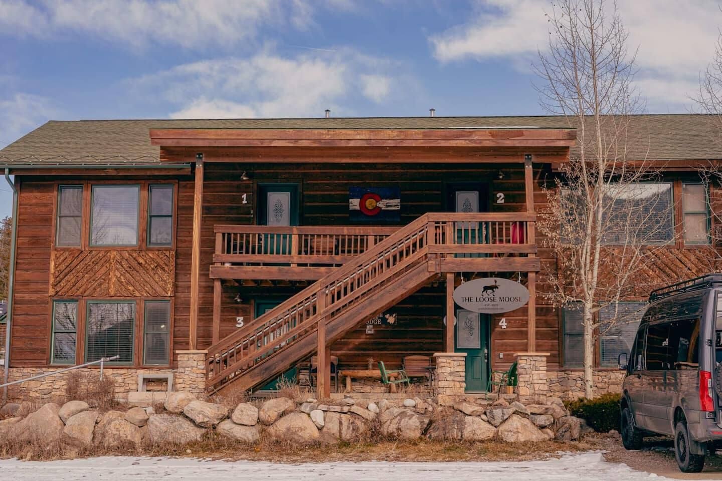 Two-story log cabin with wooden stairs, porch, and a Colorado flag. A van is parked on the right.