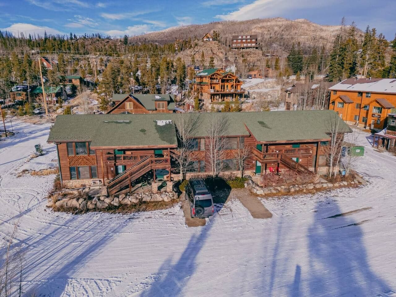 Lodge-style buildings in a snowy landscape, with a vehicle parked out front and mountain in the background.