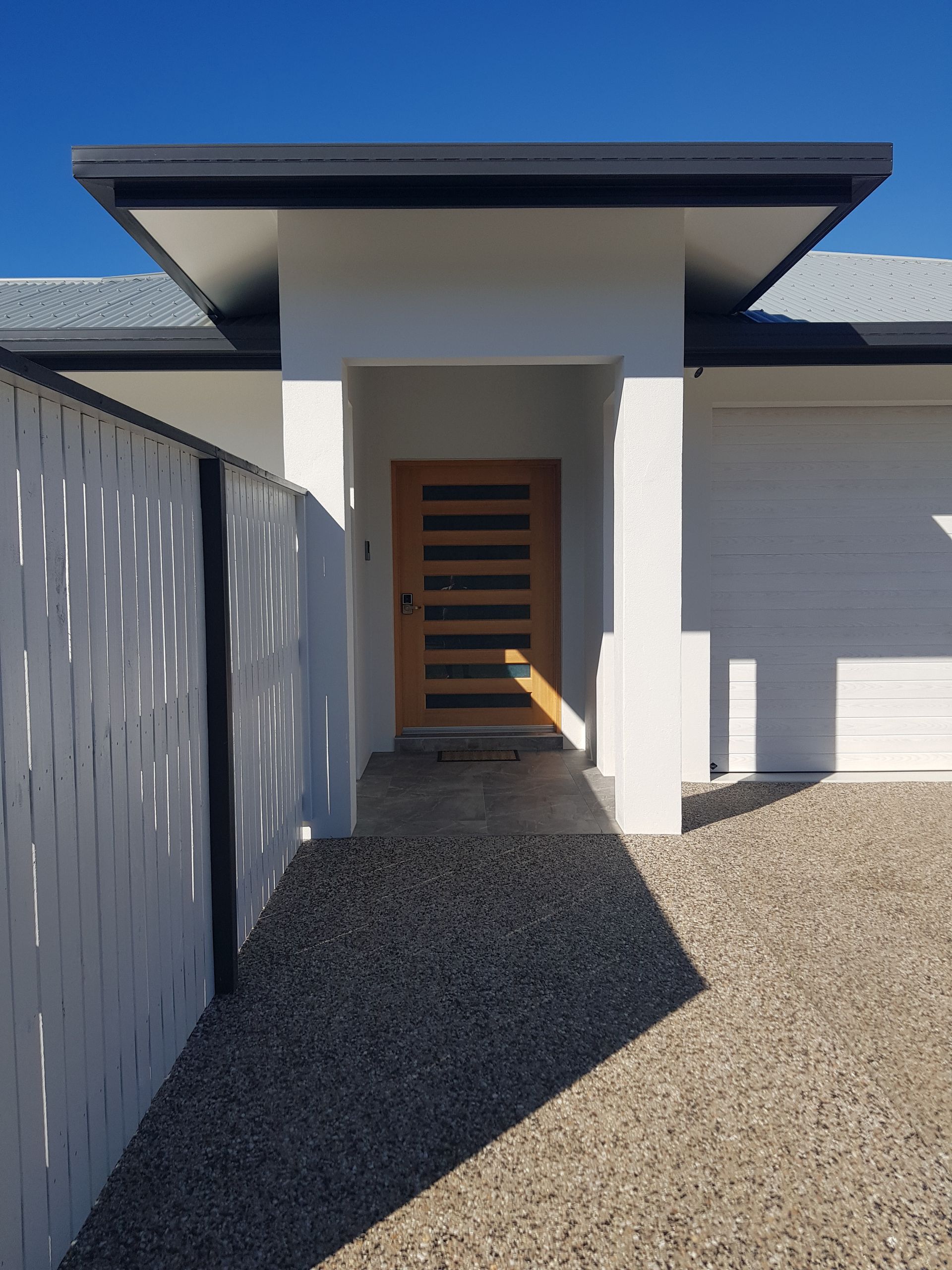 Entrance to a modern white house with light brown wooden door under a grey roof, with a gravel path and white fence — Construction Company in Townsville, QLD