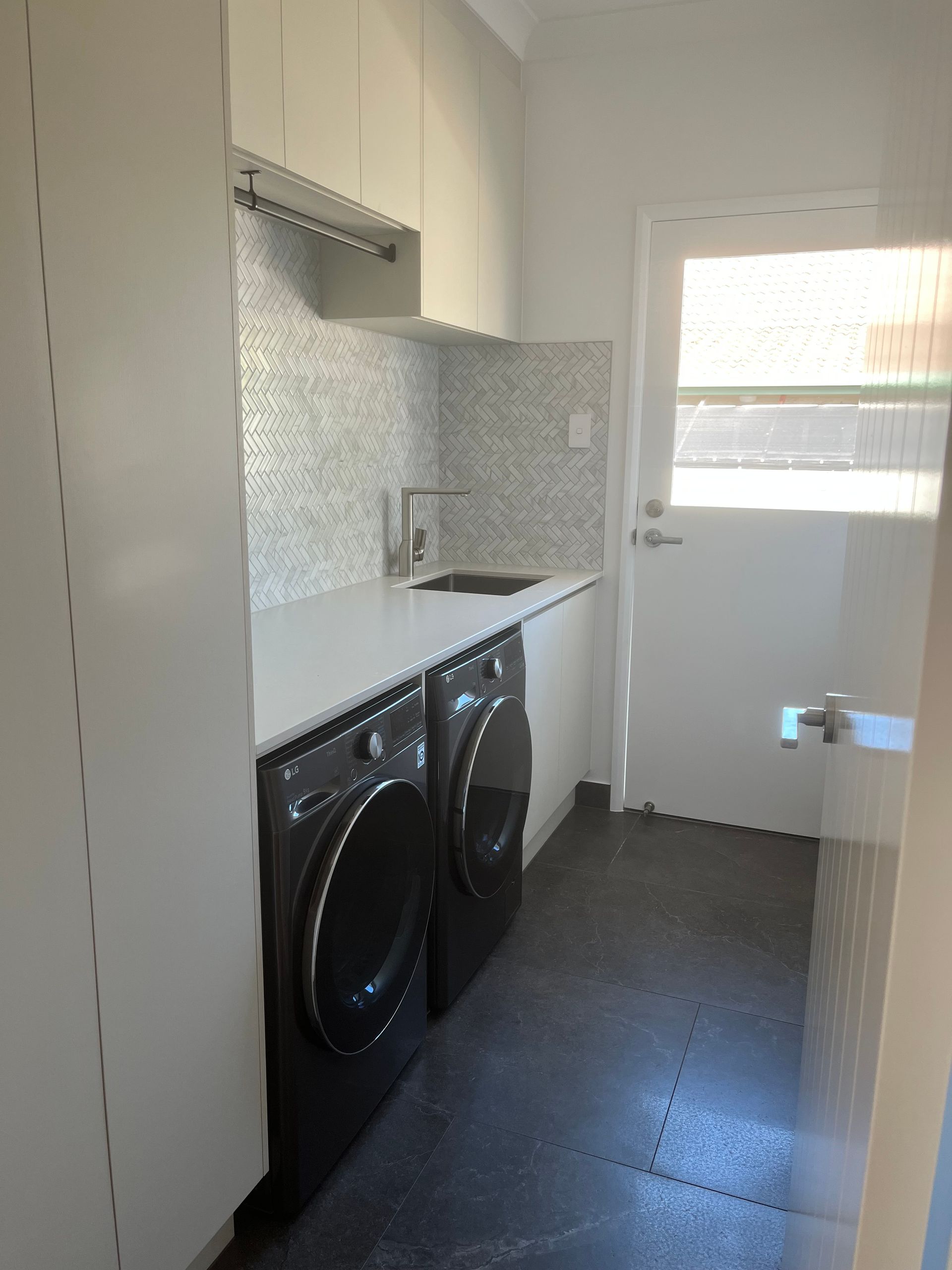 Laundry room with white cabinets, dark gray floor, two black washing machines, and a door — Construction Company in Townsville, QLD