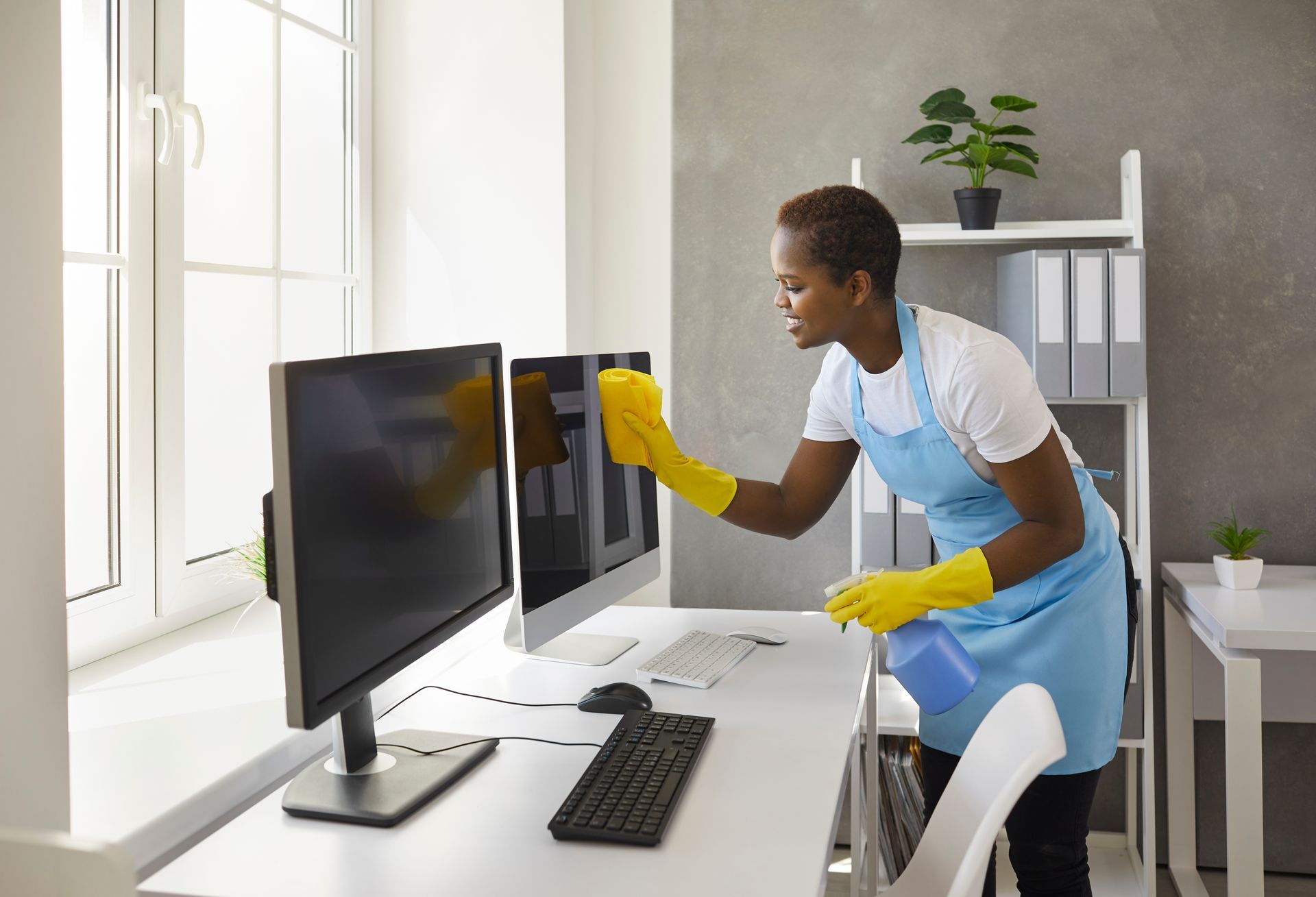 A woman is cleaning two computer monitors in an office.