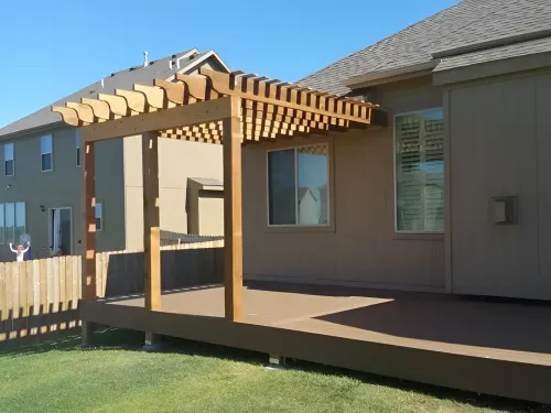 Wooden pergola on a brown deck attached to a tan house; sunny day.