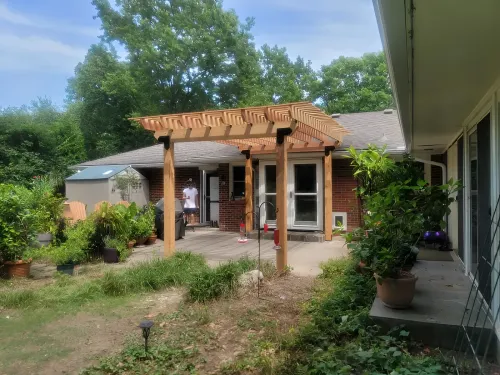 A wooden pergola covers a patio, attached to a brick house. Plants and a person stand in the yard.