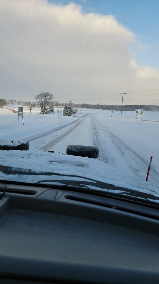 Snow-covered road viewed from a vehicle, with tire tracks and a cloudy winter sky.