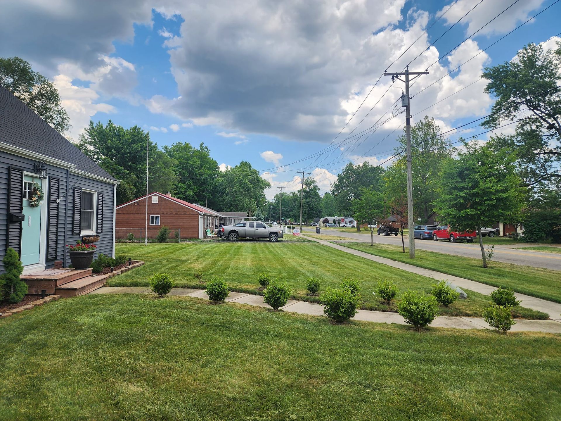 Suburban front yard with house, lawn, shrubs, trees, and a partly cloudy sky along a road