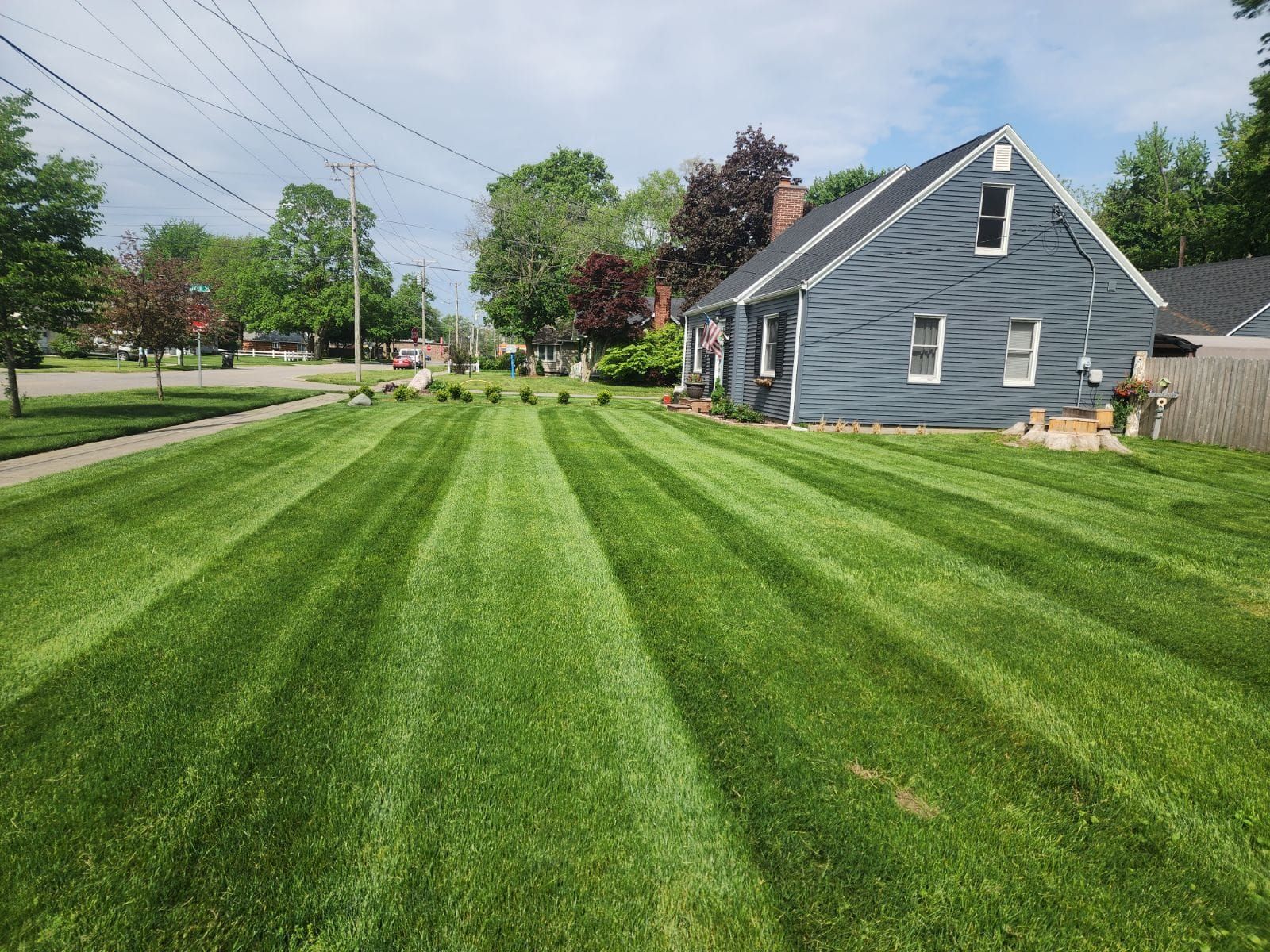 Freshly mowed green lawn beside a blue-gray house on a suburban street under a partly cloudy sky
