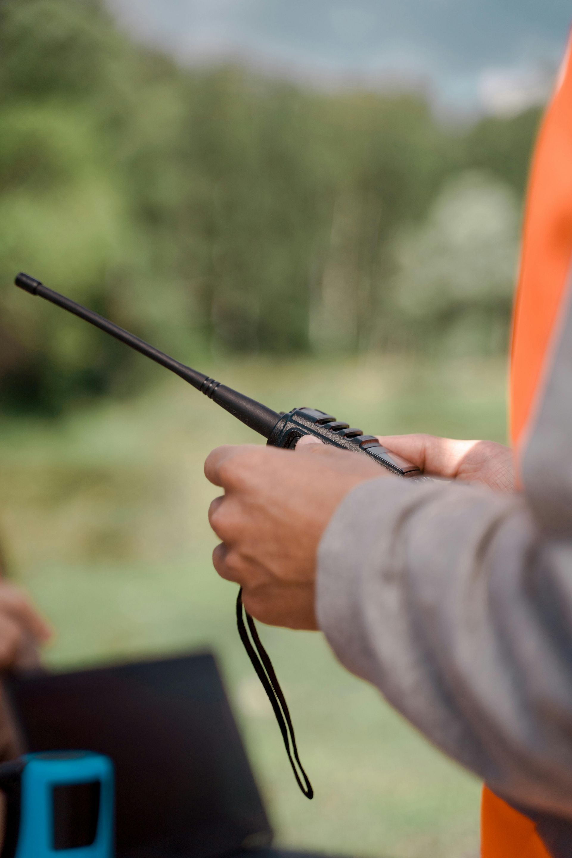 Person holding a black handheld radio with a strap outdoors, with a blurred green background