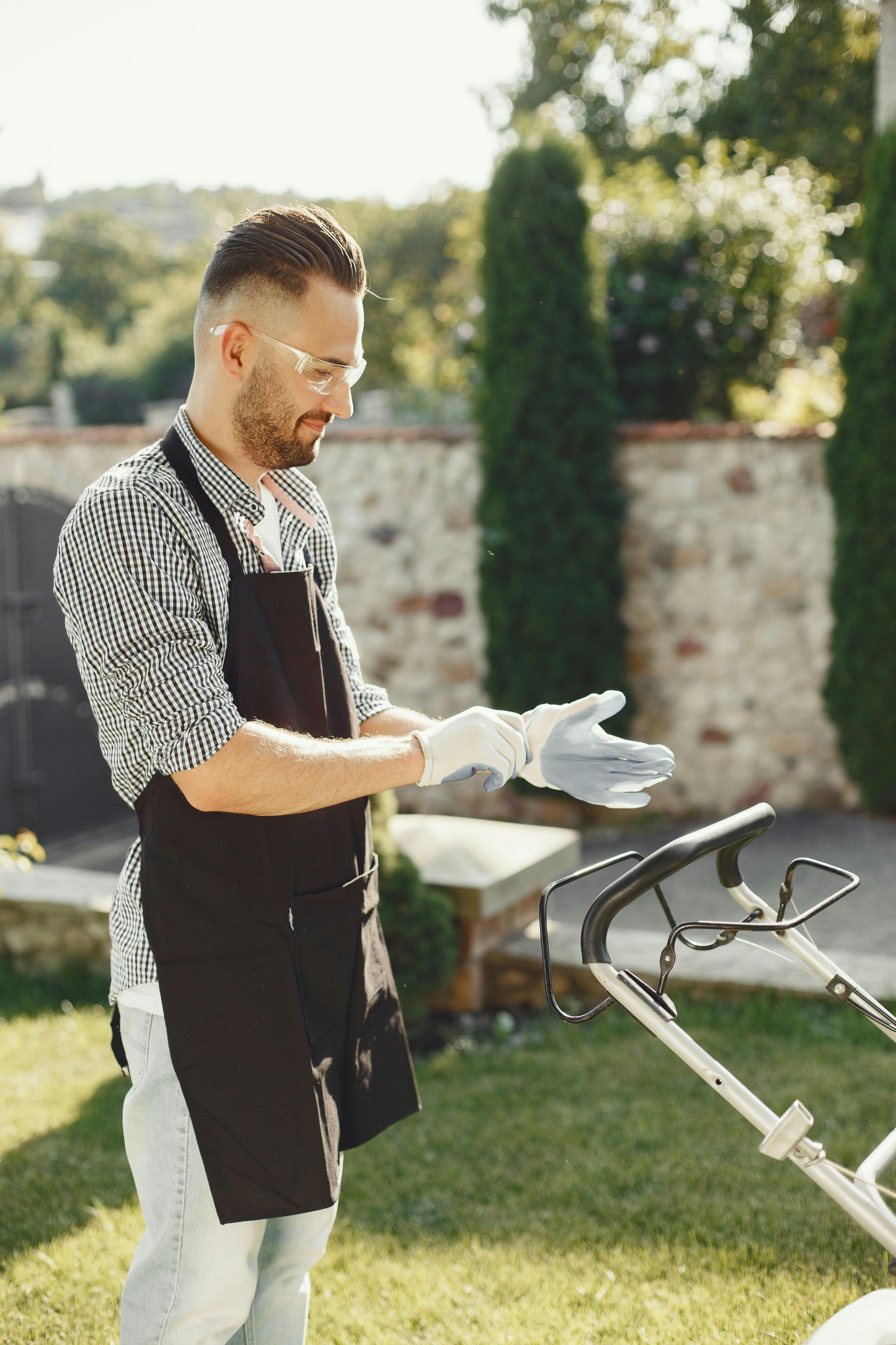 Gardener in apron and gloves inspecting a lawn mower on a sunny grassy lawn