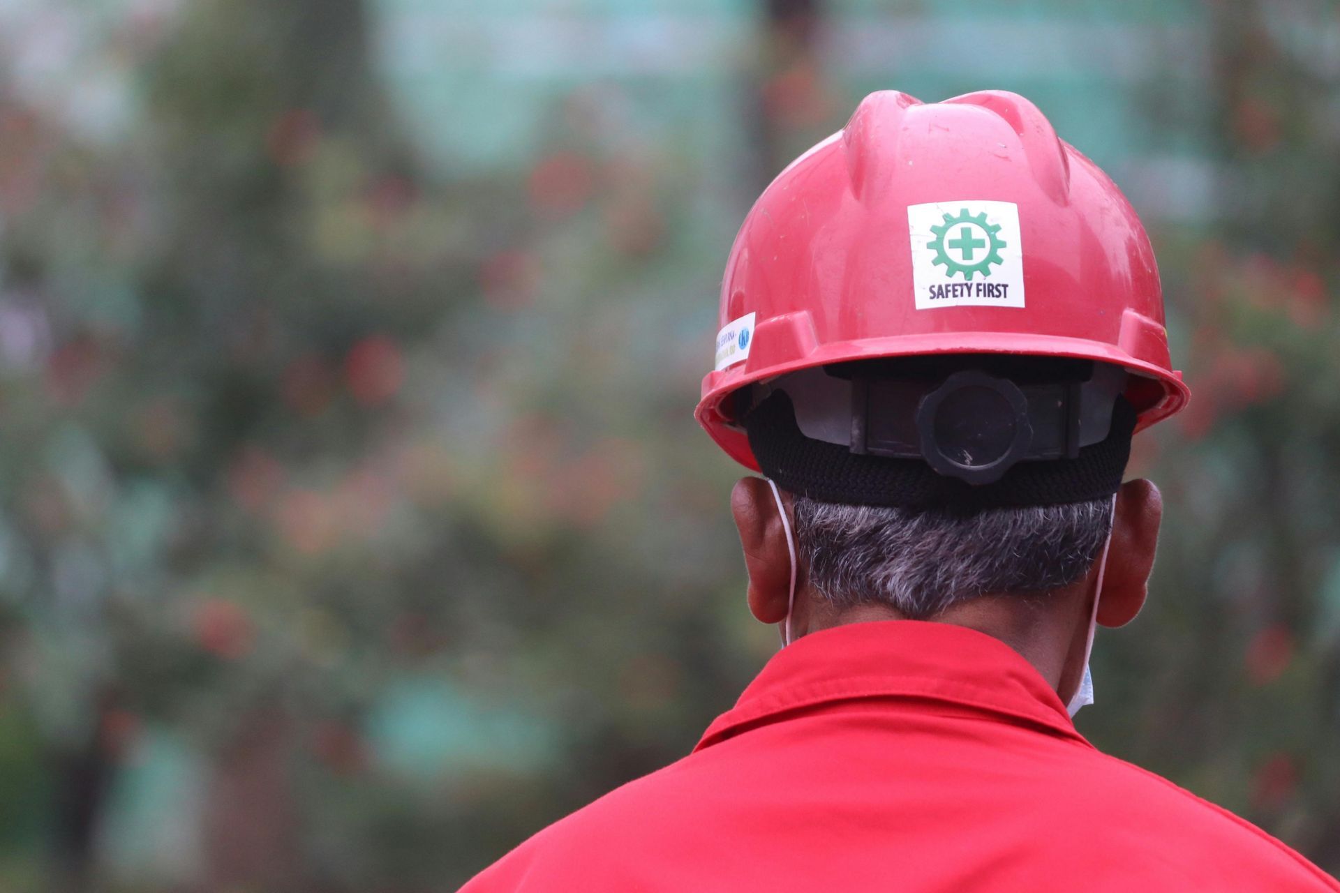Worker in a red hard hat and jacket, seen from behind against a blurred outdoor background.