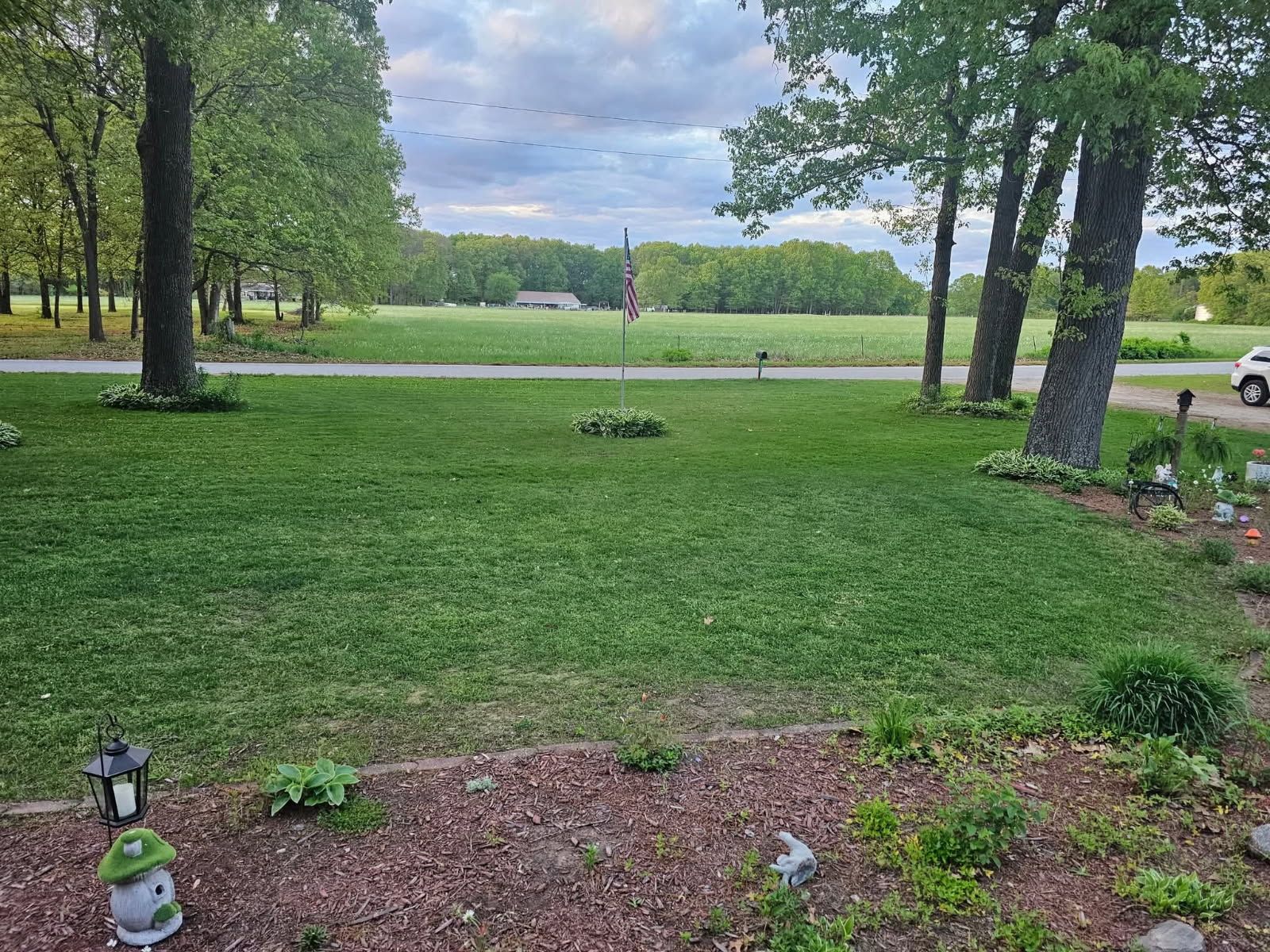 Grassy park with trees, a small road, and garden decor in the foreground under a cloudy sky