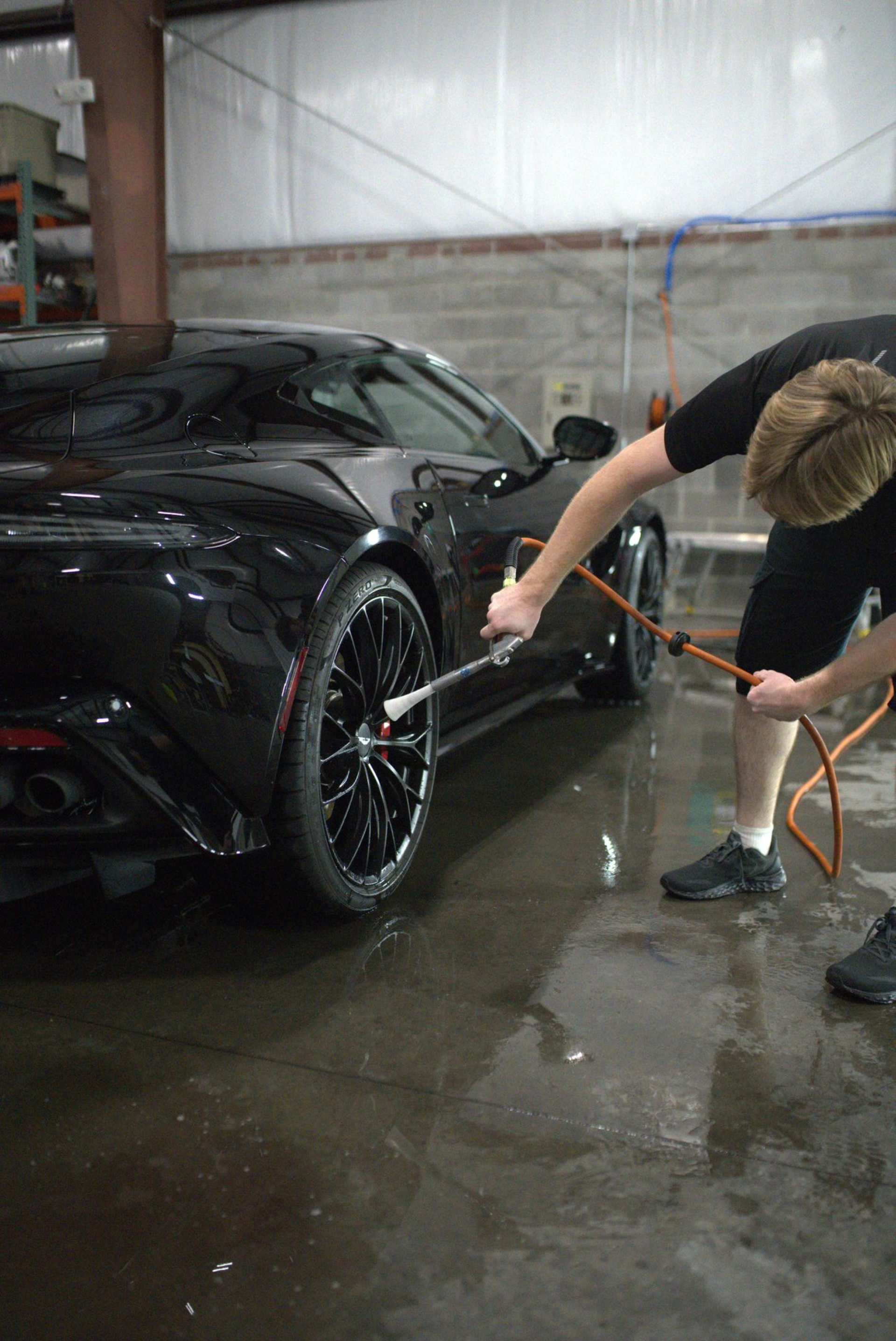 Person washing a black sports car's tire with a hose in a car wash bay.