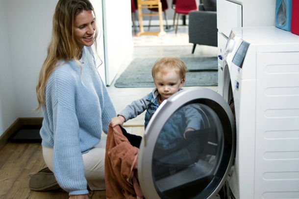 A woman and a baby are sitting on the floor loading clothes into a washing machine.