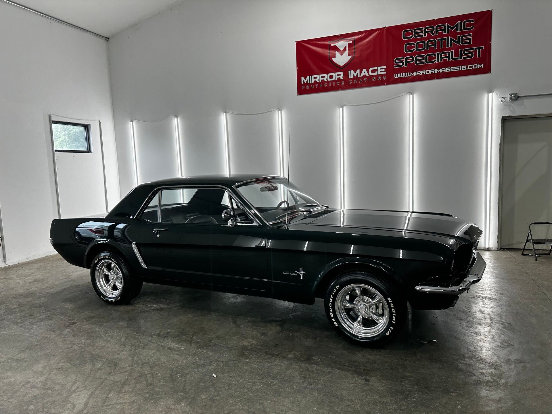 A black classic Ford Mustang coupe parked inside a well-lit showroom with a white banner on the wall.