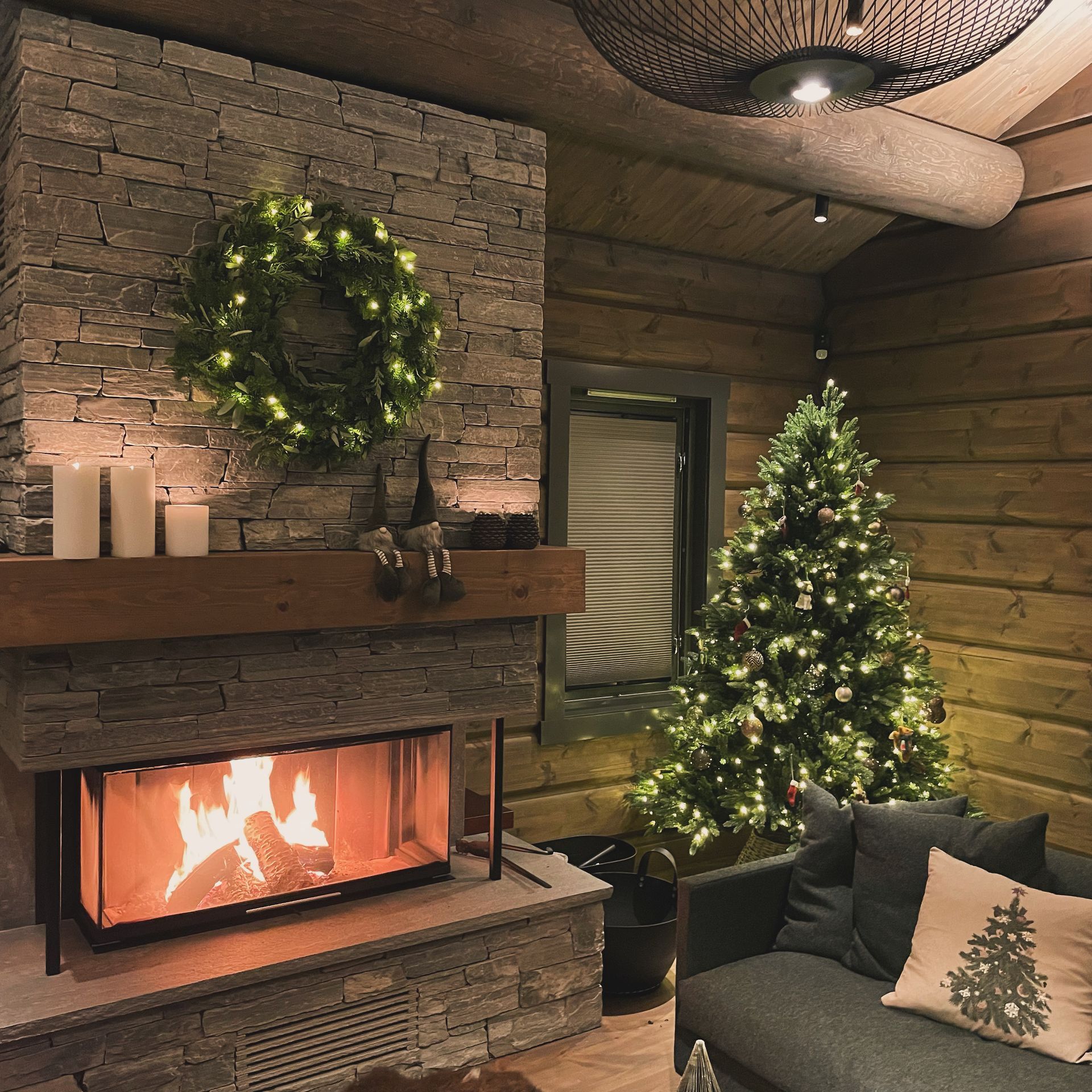 Cozy fireplace with a lit Christmas tree and wreath in a log cabin interior.