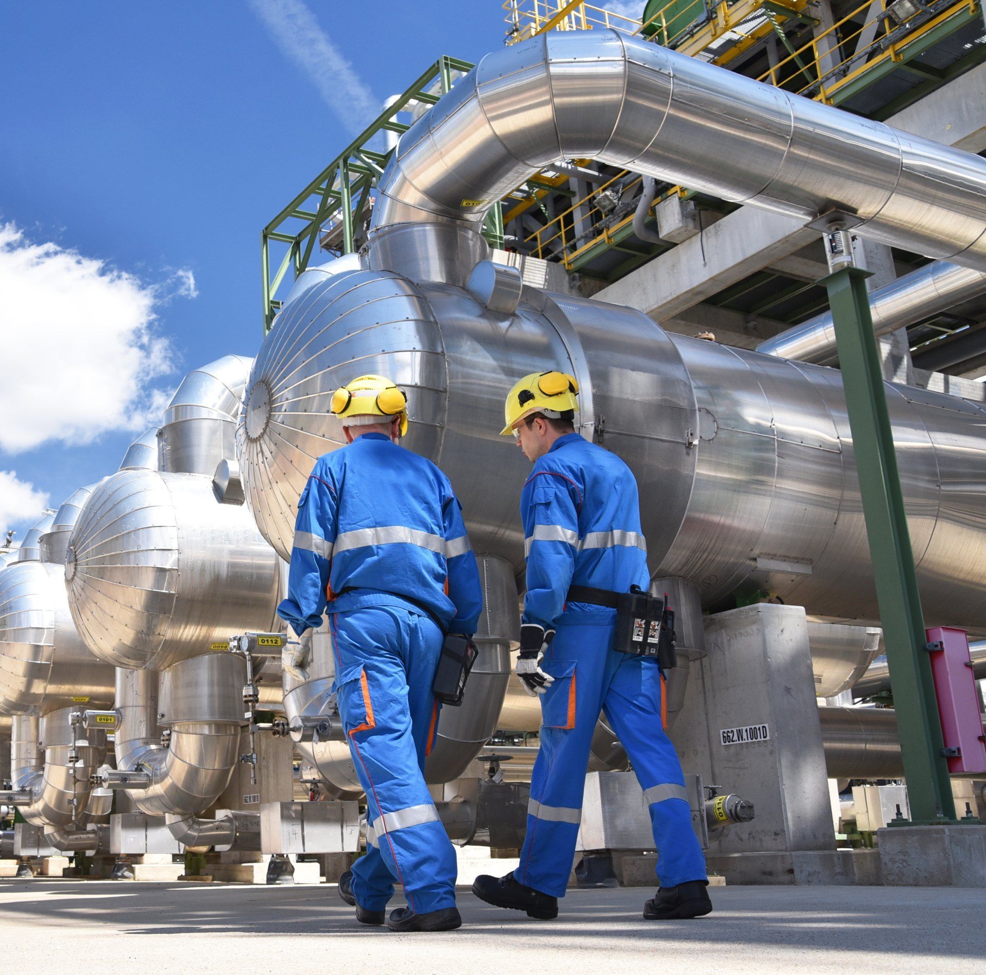 Two men in blue jumpsuits are walking in front of pipes