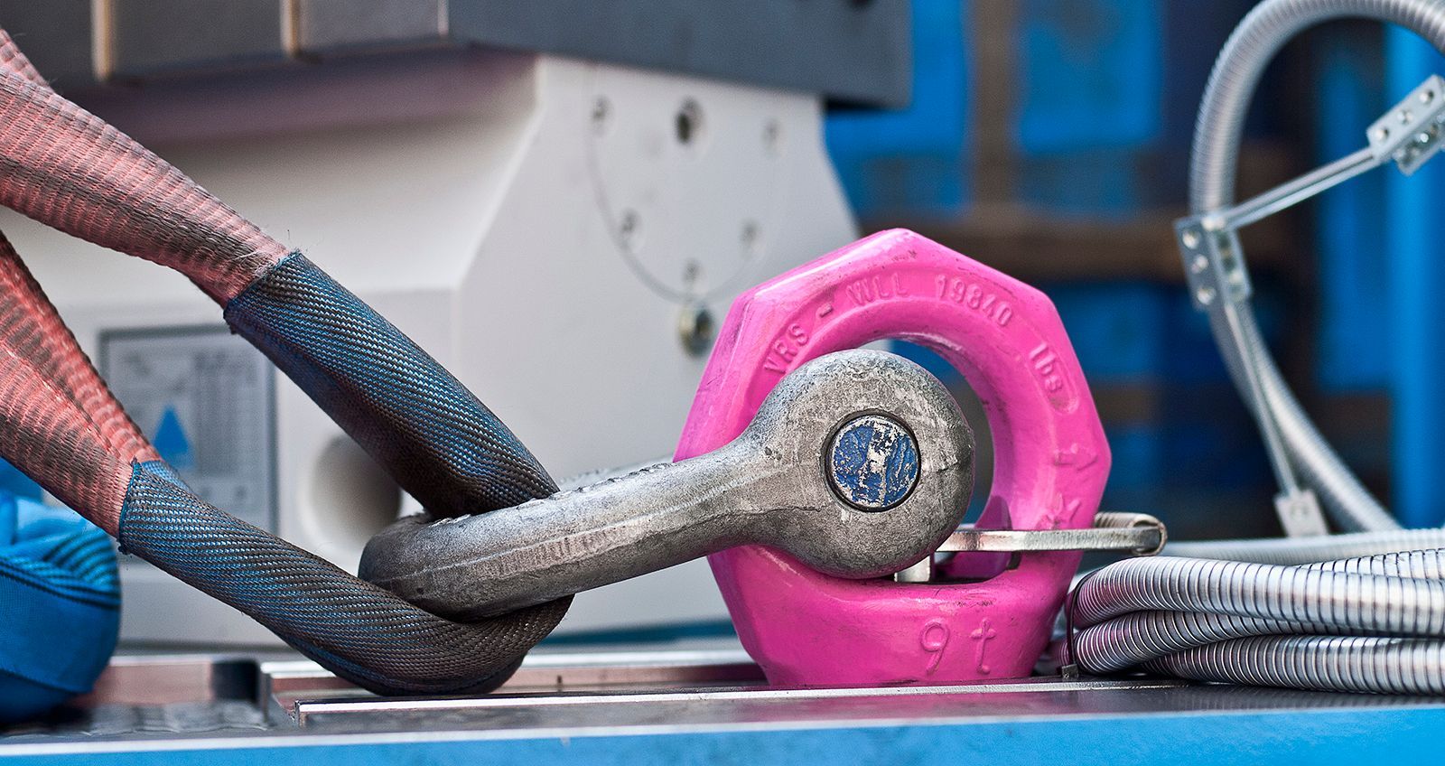 Grillete rosa conecta una eslinga textil azul a un cable metálico, entorno industrial.