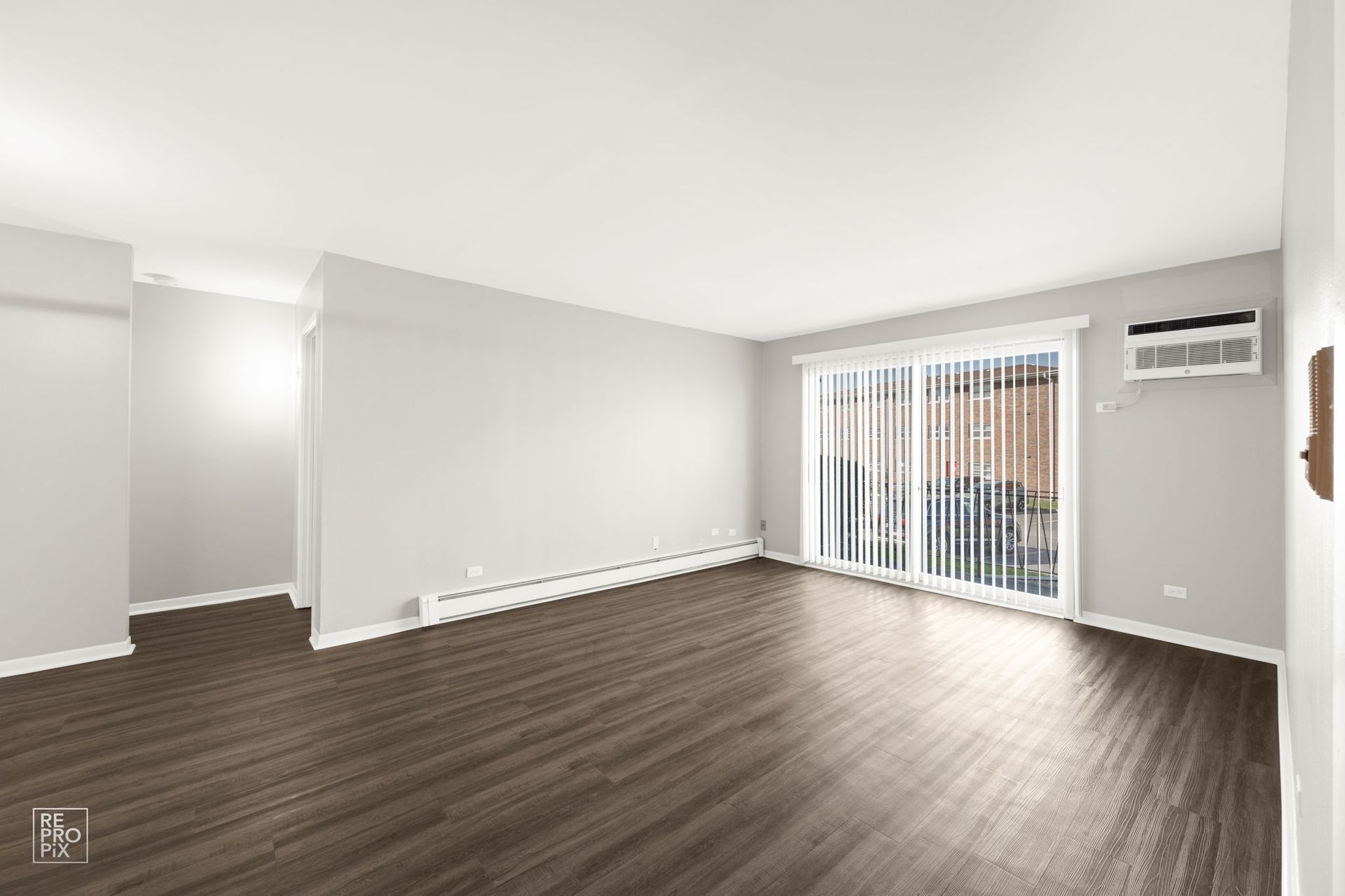 Empty apartment living room with dark wood floors, sliding glass door, and air conditioner.