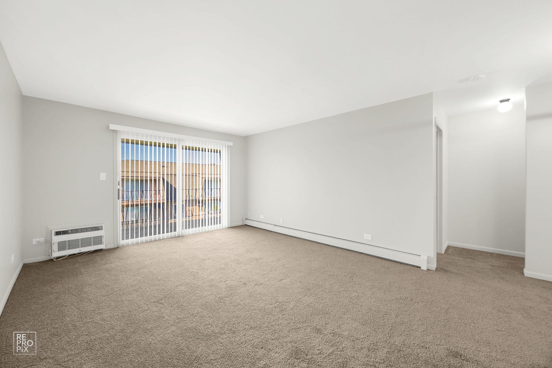 Empty apartment living room with sliding glass door, beige carpet, and light gray walls.