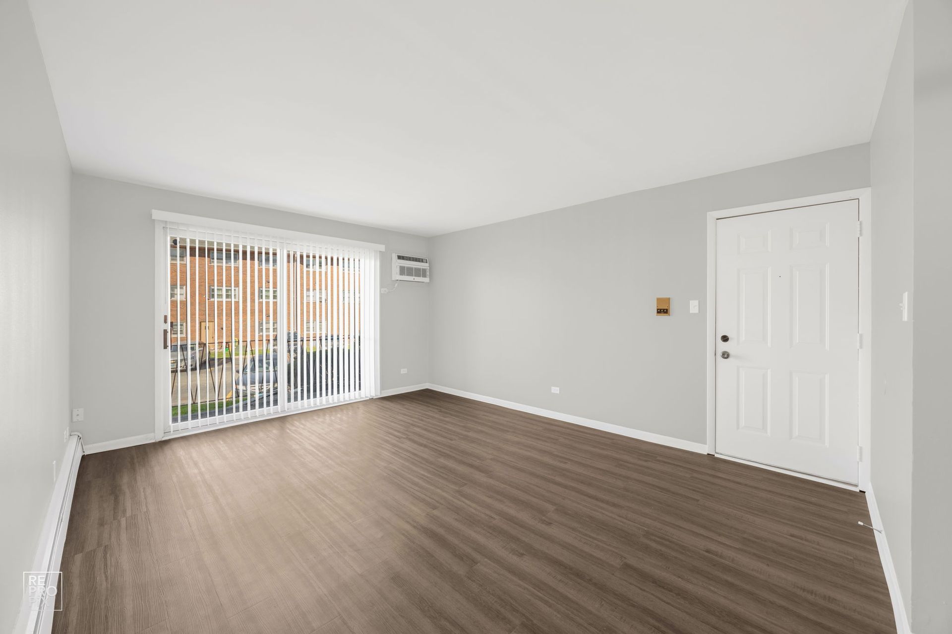 Empty living room with dark wood-look flooring, sliding glass door, white door, and gray walls.
