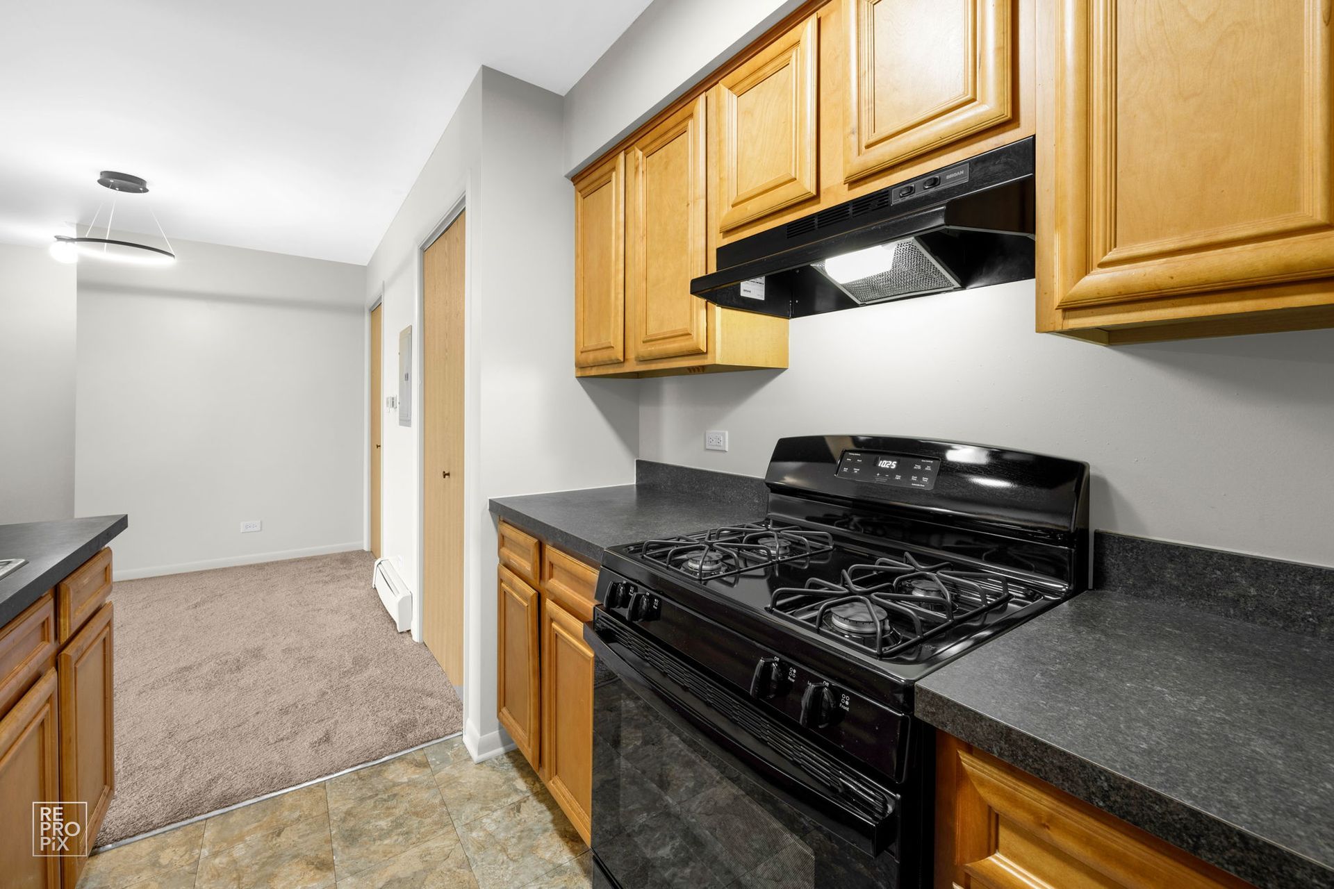 Kitchen with light brown cabinets, black appliances, and gray countertops. Hallway in background.