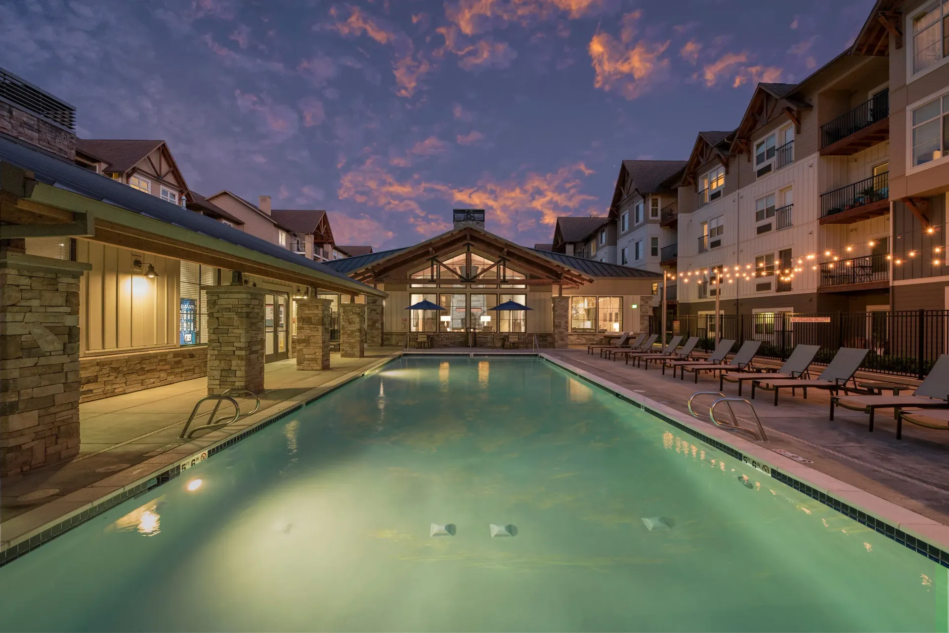 Outdoor pool area at a multifamily community with lounge chairs and string lights at dusk.