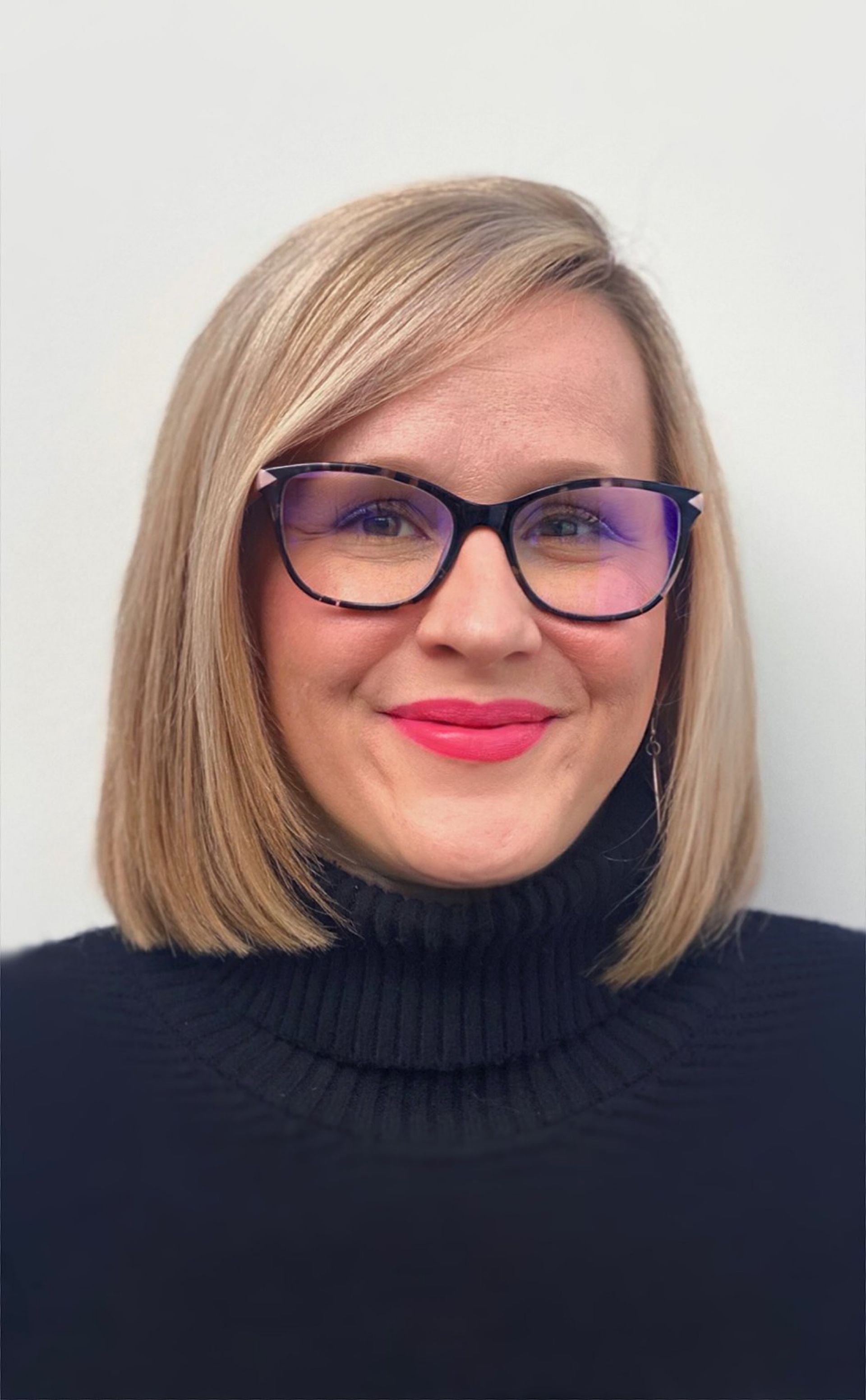 Woman with blonde bob and glasses wearing a black turtleneck smiles in front of a white wall.