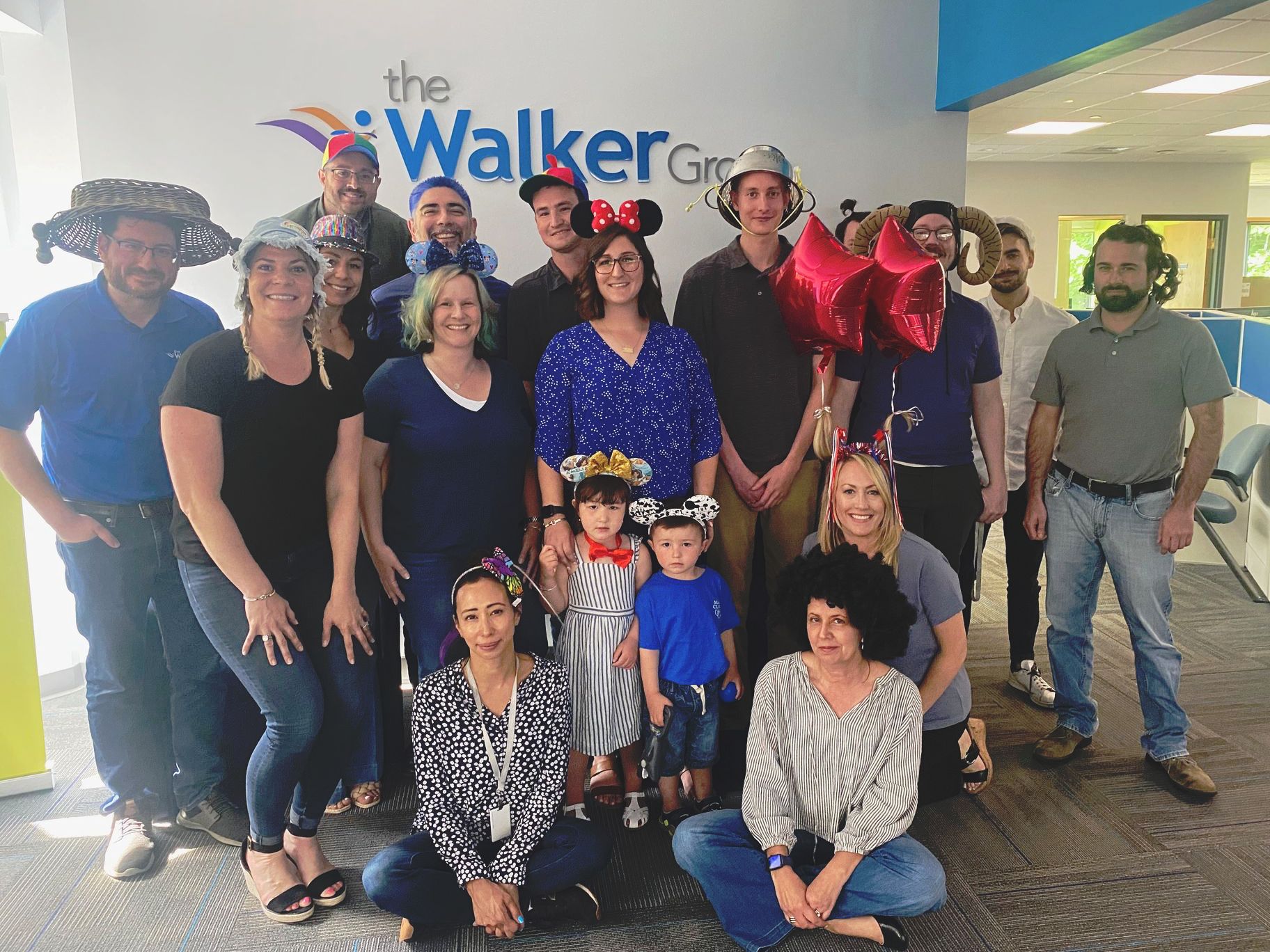 Group of people wearing novelty hats pose in an office setting in front of the Walker Group logo.