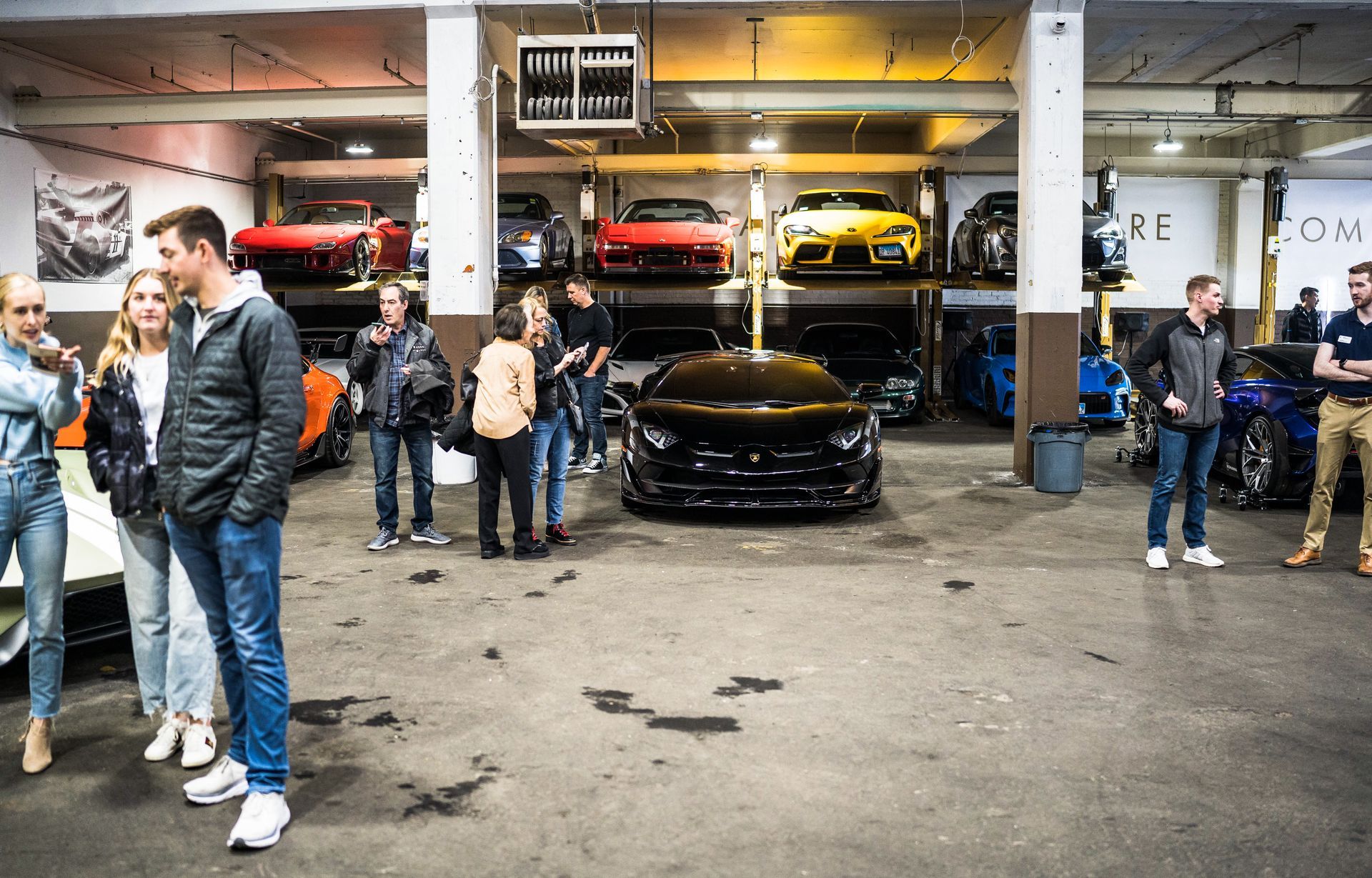 People viewing cars in a garage; some cars are on lifts, others are on the floor.