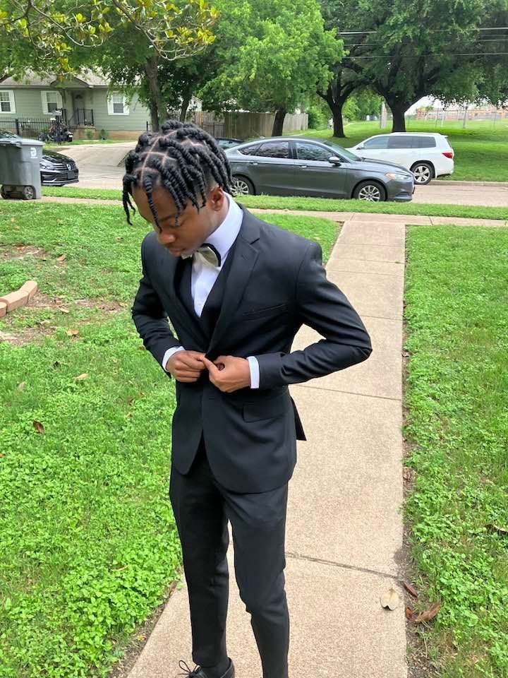 A young man in a black suit and bow tie is standing on a sidewalk.