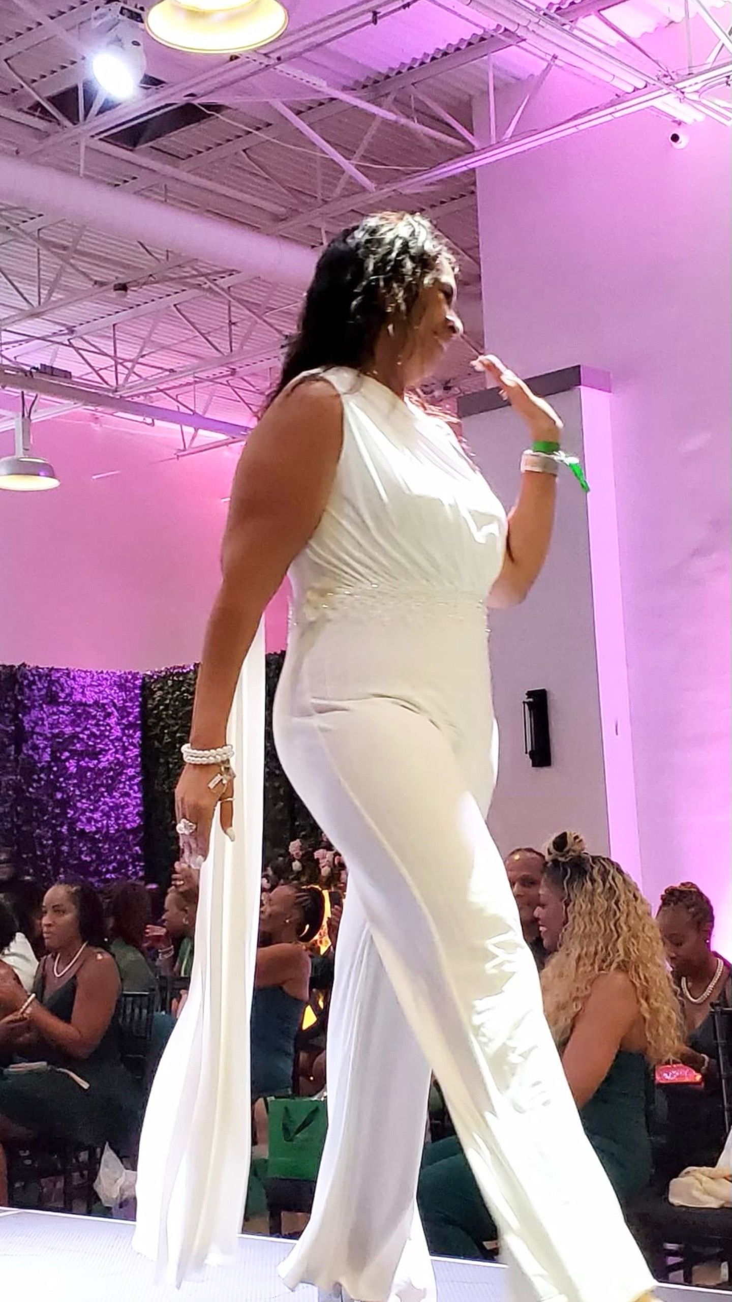 A woman in a white jumpsuit is walking down a runway at a fashion show.