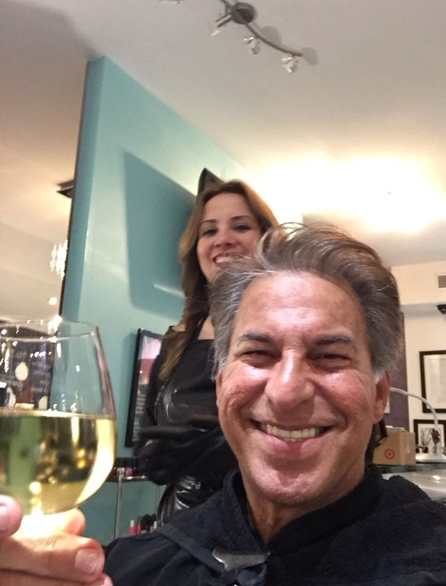 Man holding wine glass, smiling in a salon. Woman in background. Bright lighting, teal wall.