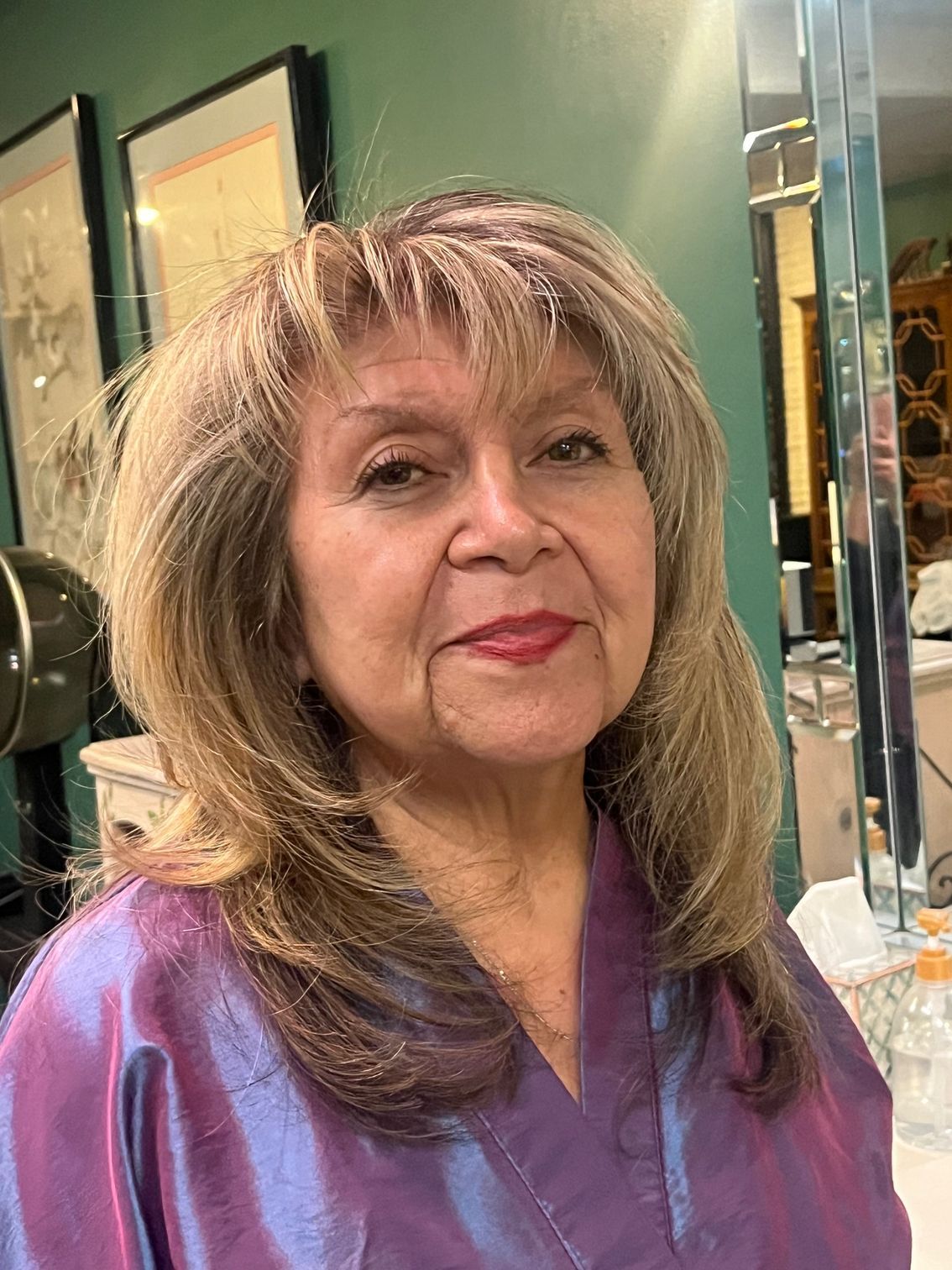 Woman in a salon wearing a purple smock, with layered blonde hair.