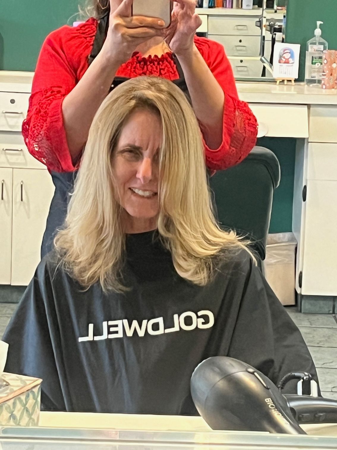 Woman with blonde hair in a salon chair, wearing a black cape. Hairdresser in red shirt holds phone up, taking a photo.