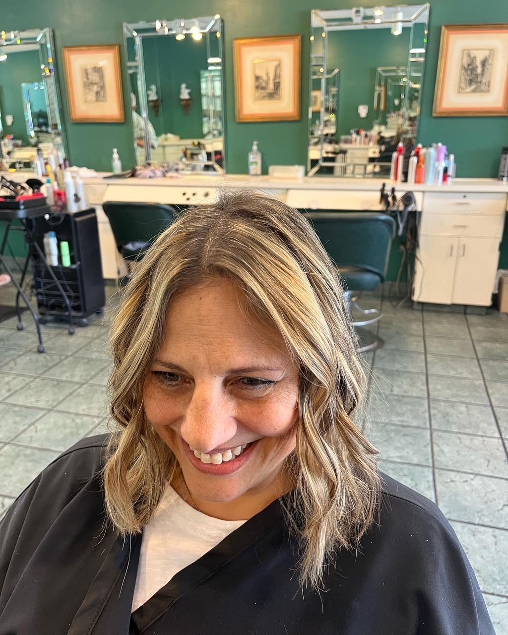 Woman with wavy blonde highlights at a salon, smiling. Green walls, mirrors and styling stations are in the background.