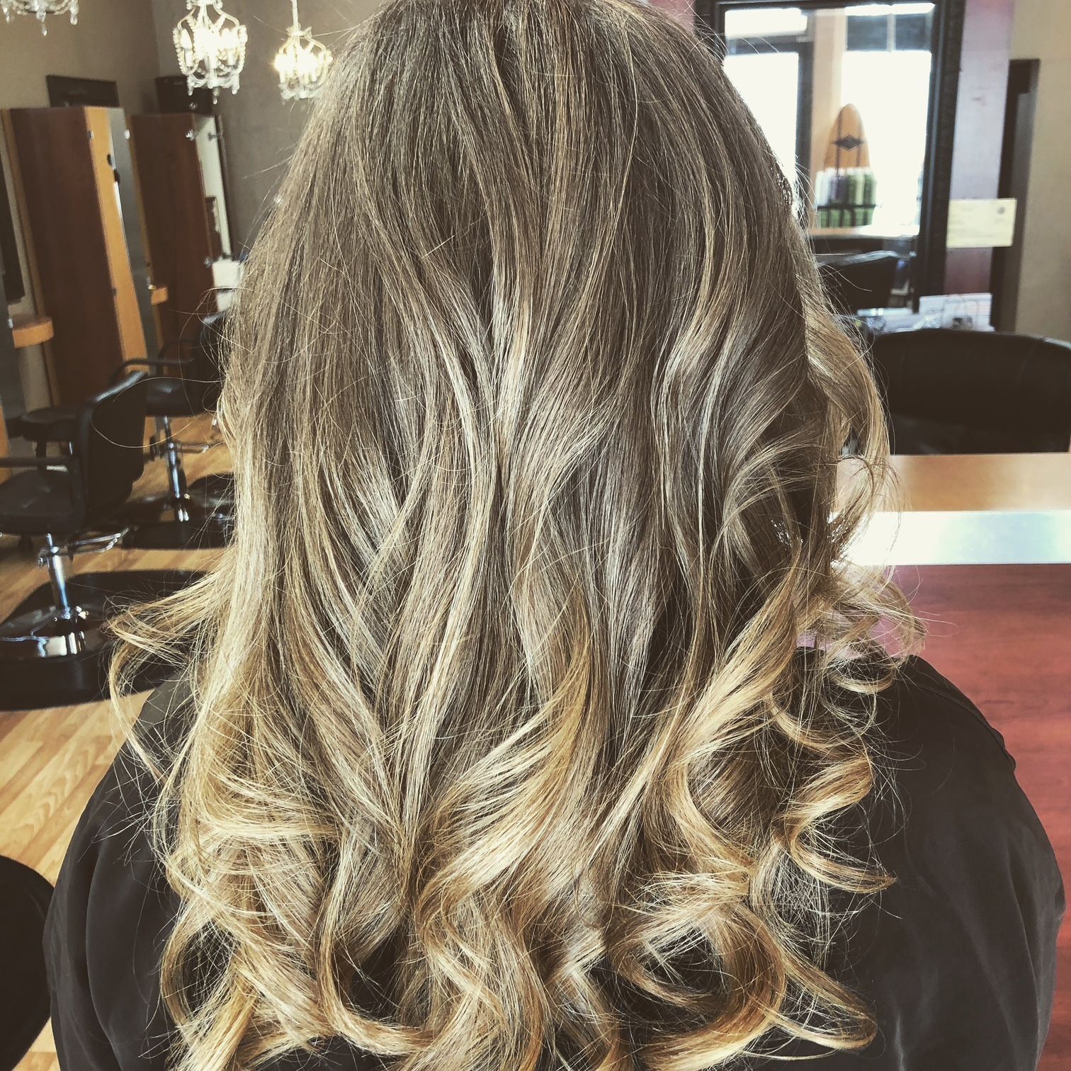 Woman's back with long, wavy hair. It transitions from brown to blonde, styled in curls, salon setting.