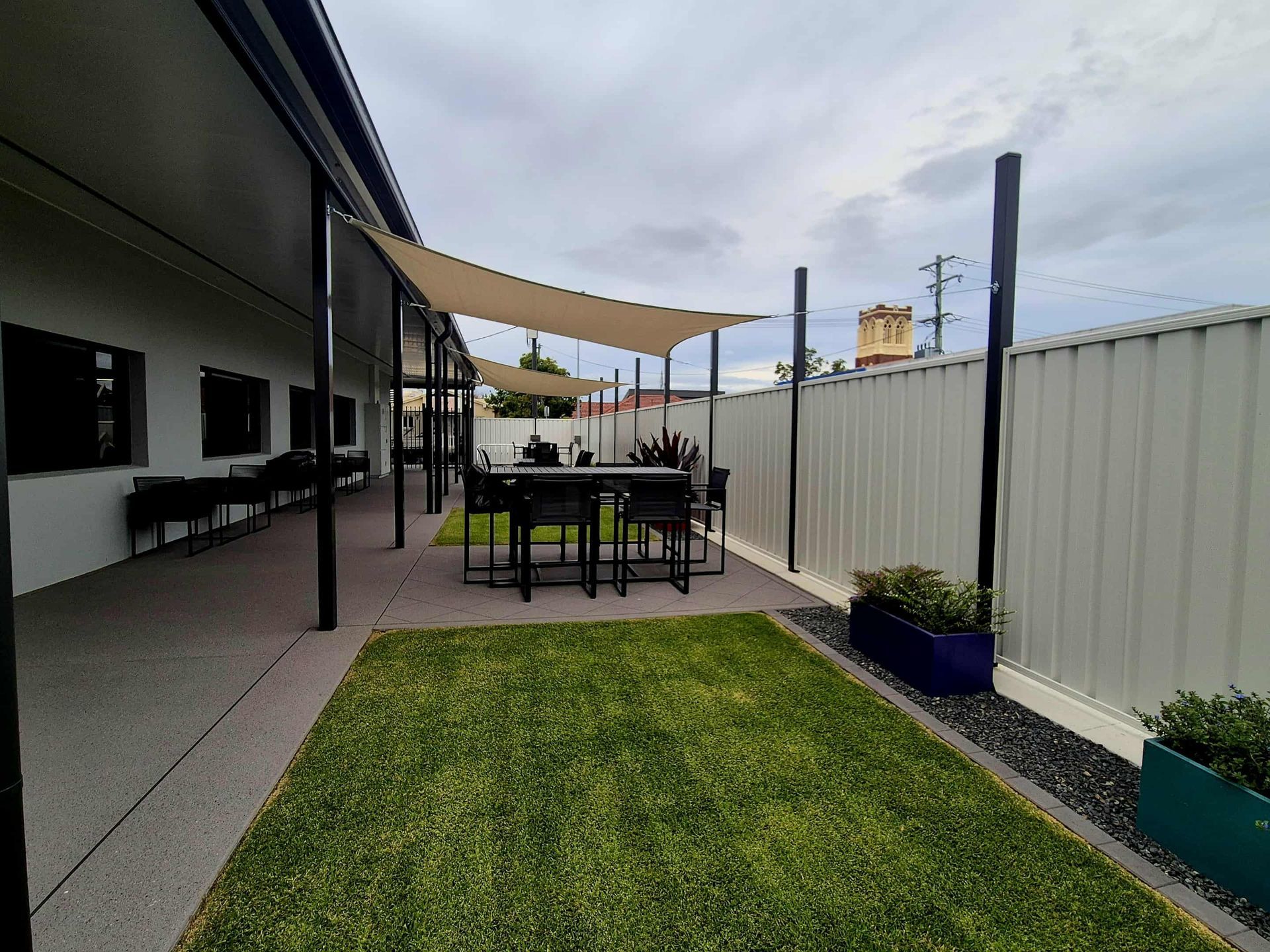 Outdoor Patio With a Table and Chairs Shaded by a Tan Sail Against a Cloudy Sky — On The Mark Landscaping in Branyan, QLD