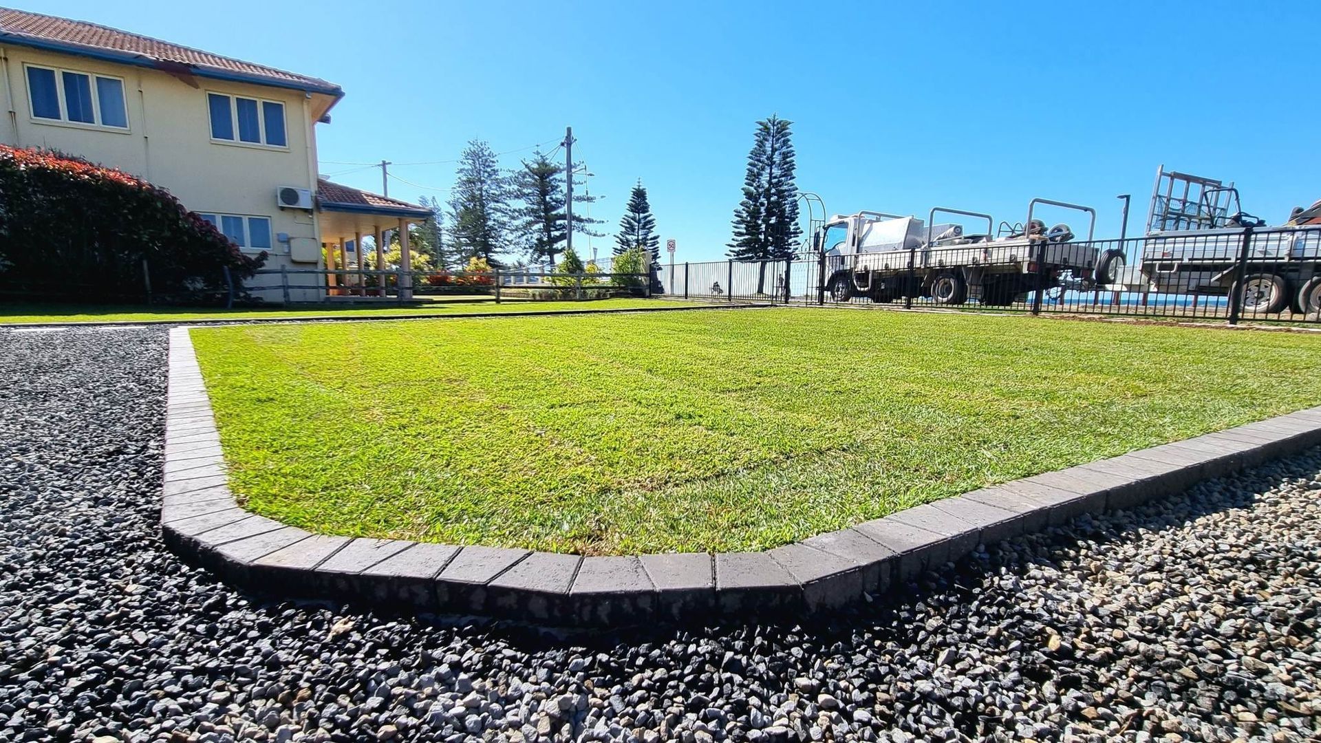 Green Lawn Bordered by Gray Pavers and Gravel, With a Building and Trucks — On The Mark Landscaping in Branyan, QLD