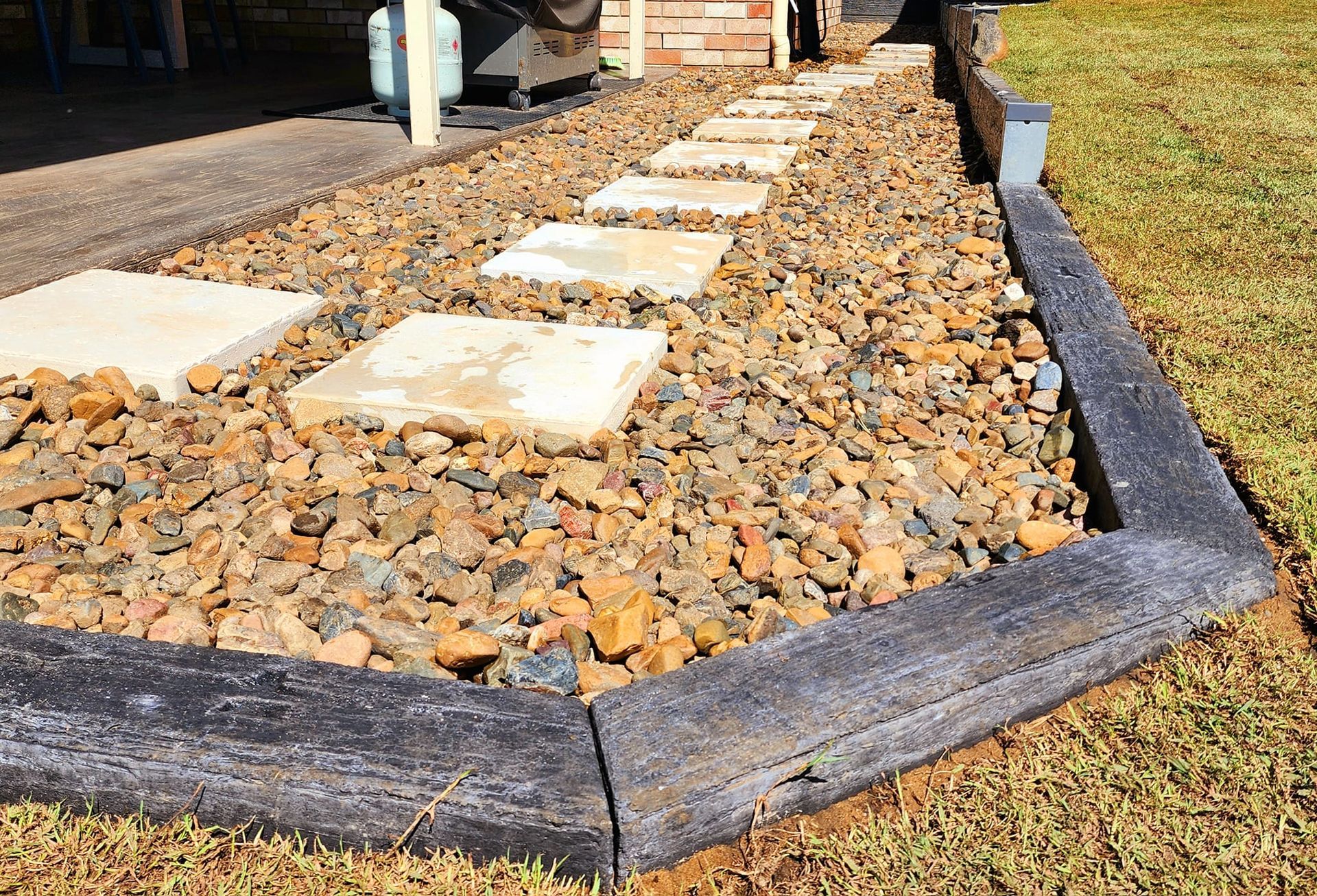 Pathway of Stepping Stones Through a Rock Garden, Bordered by Gray Wooden Edging — On The Mark Landscaping in Branyan, QLD